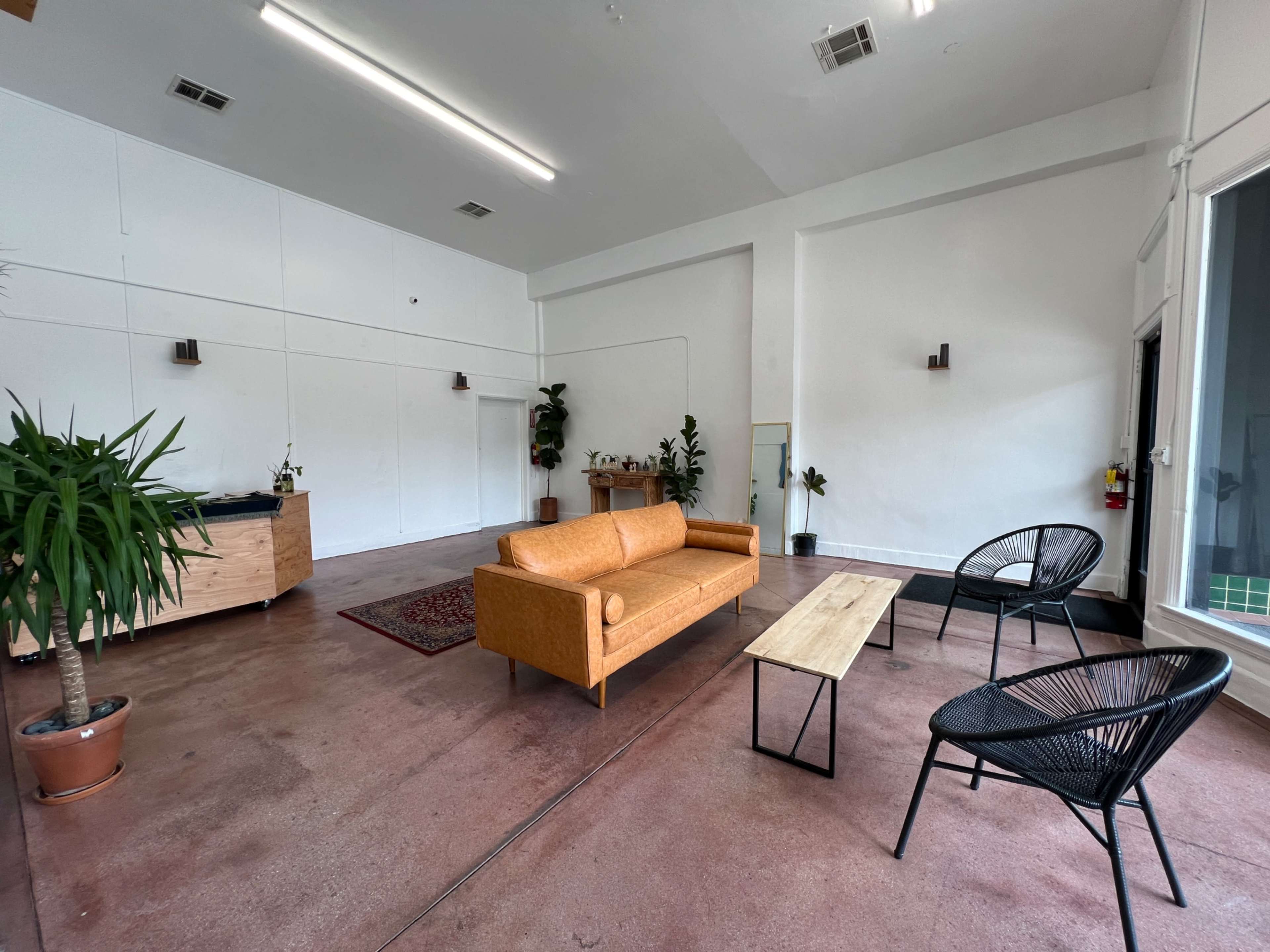 A spacious room features a tan couch, a wooden coffee table, two black chairs, and potted plants.