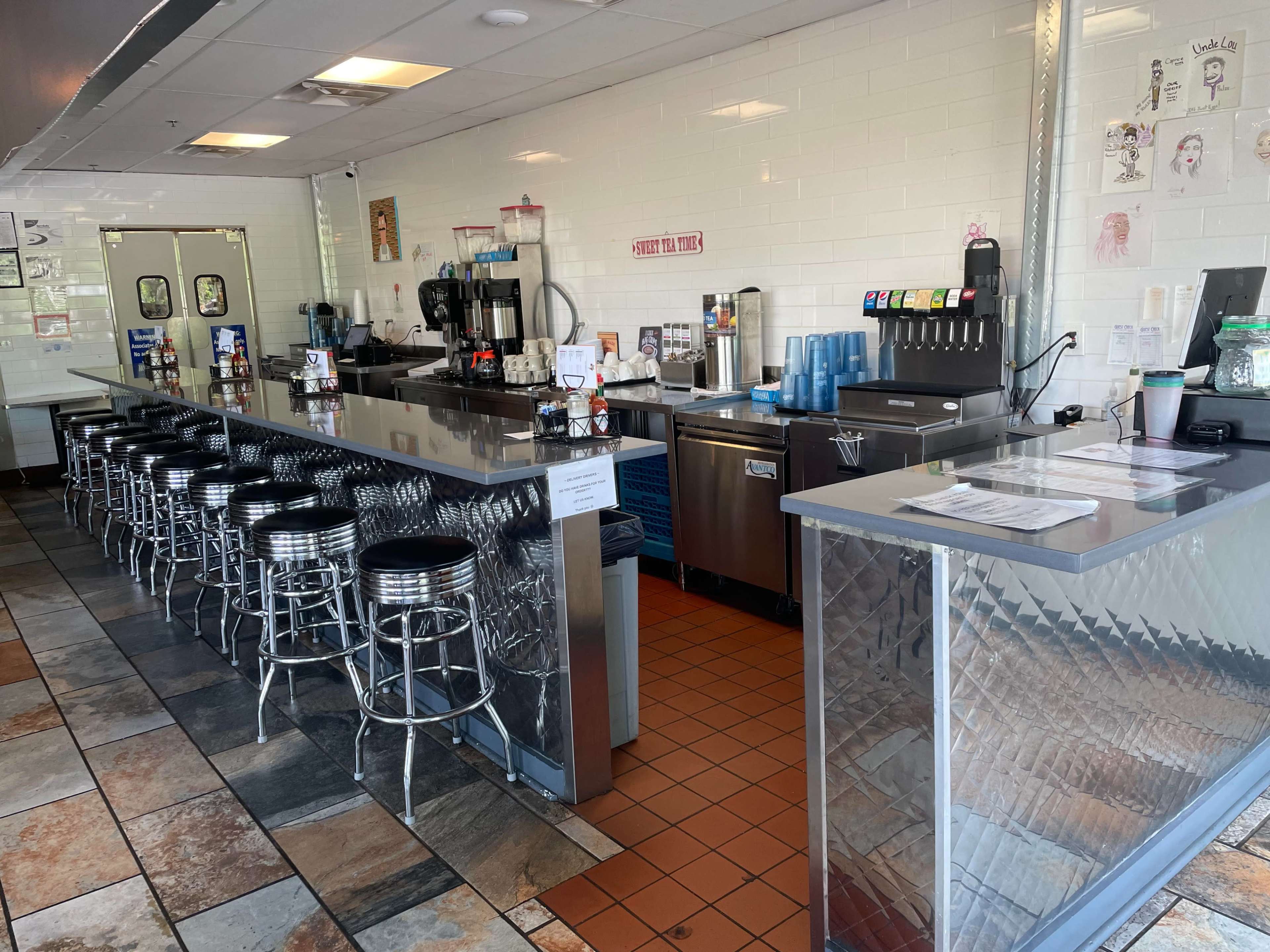 The image shows a diner-style cafe interior with a metallic counter, barstool seating, and a drink station.