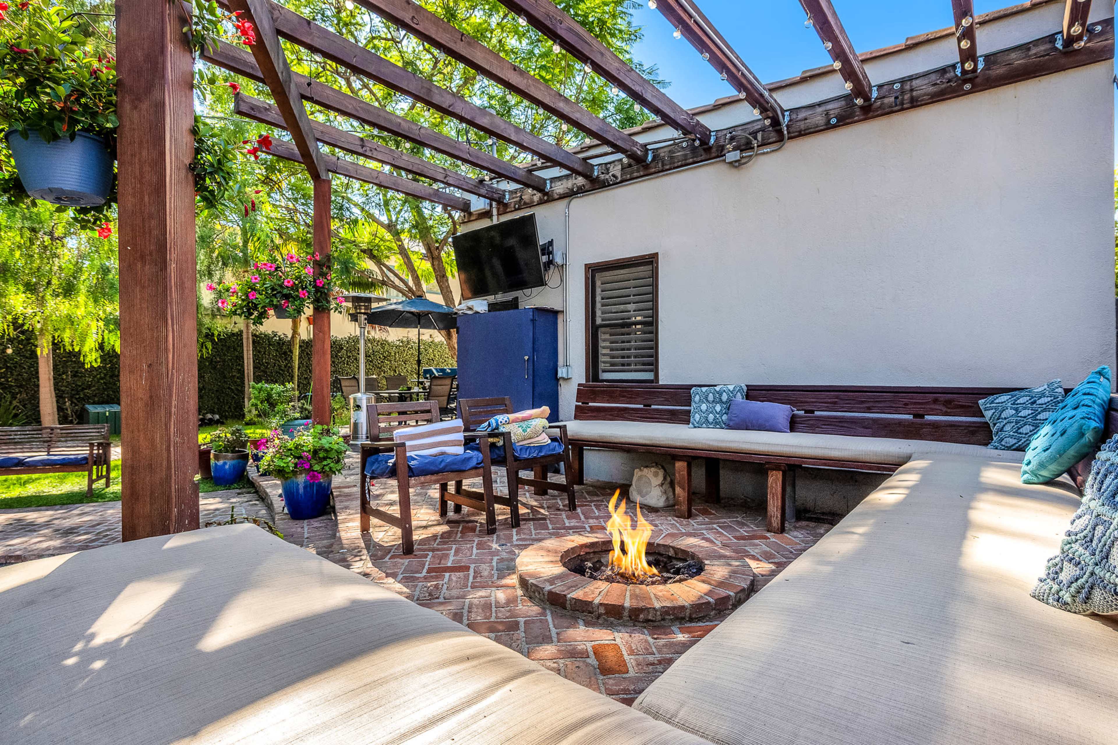 The image shows a backyard patio with a circular fire pit surrounded by a seating area and a wooden pergola, adorned with hanging plants and a television mounted on a wall.