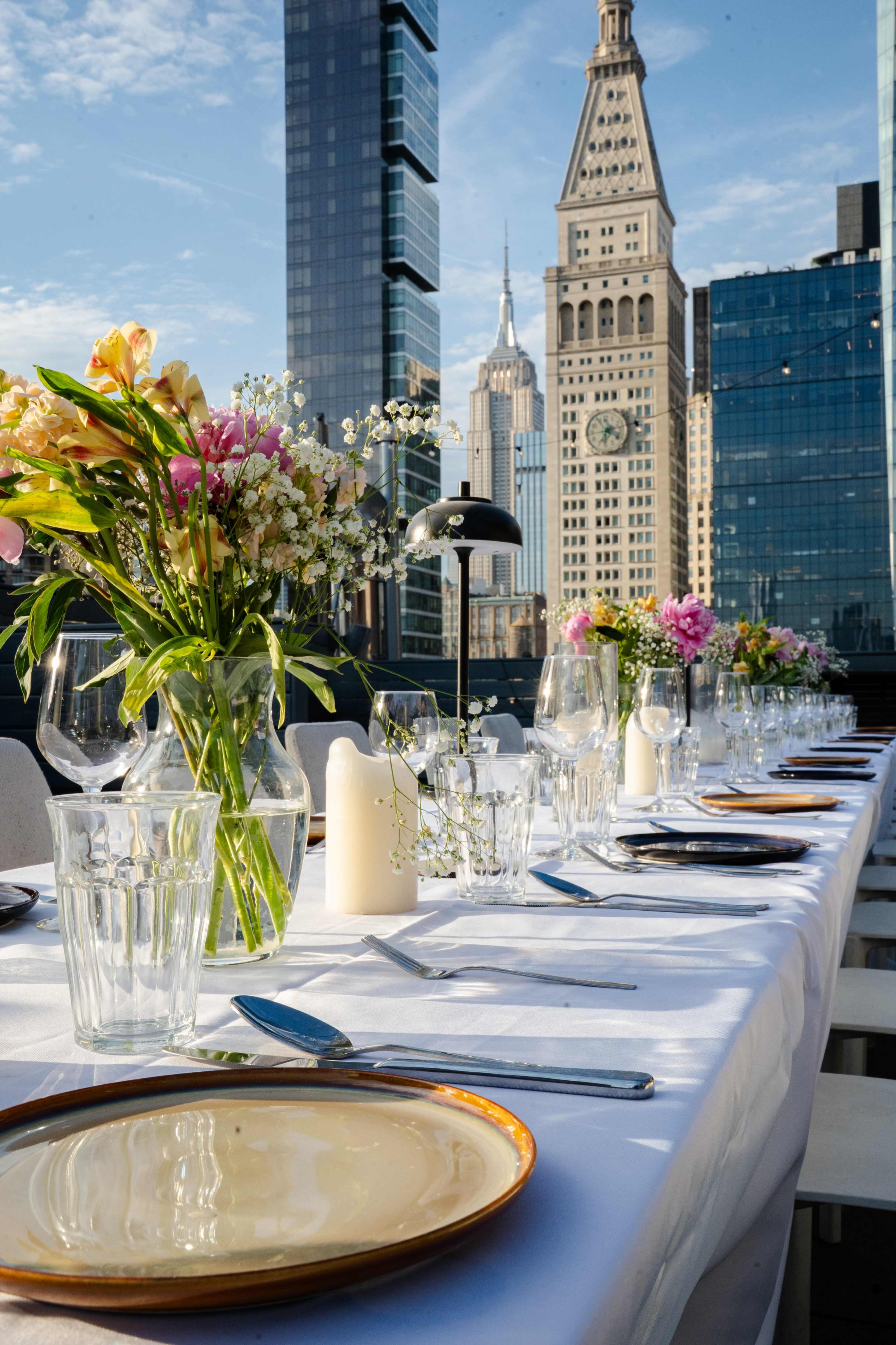 A long dining table set with plates and glassware overlooks the Empire State Building amid modern skyscrapers.