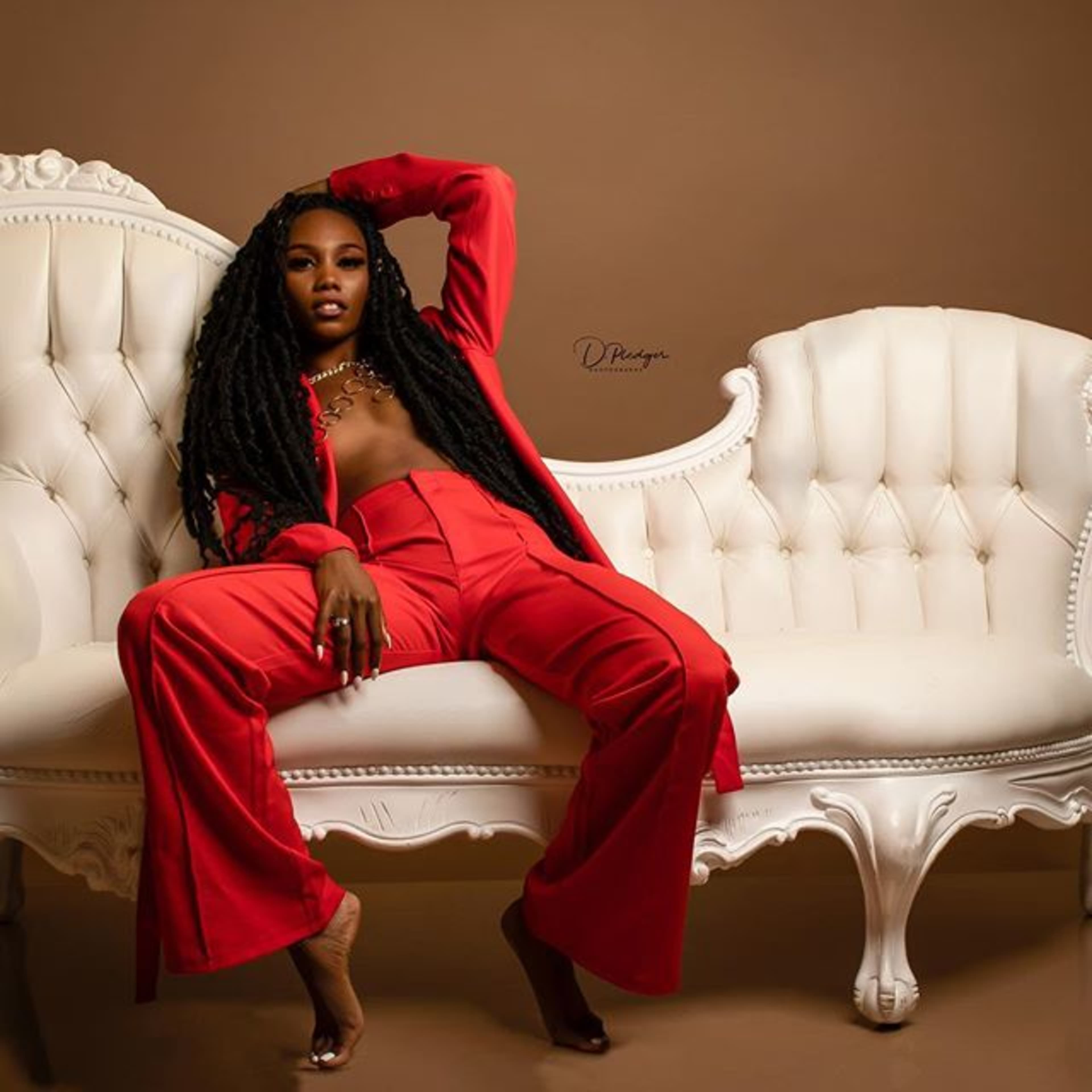A woman in a red outfit reclines on a white vintage-style sofa against a brown backdrop.