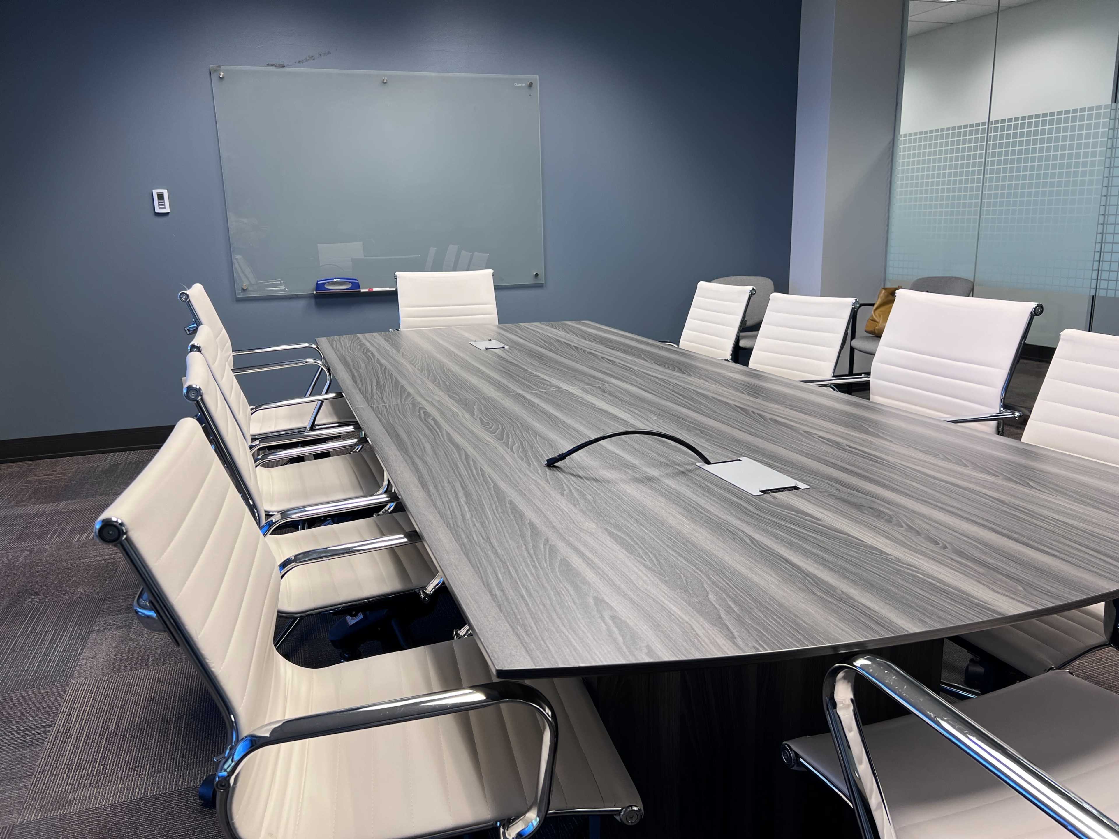 A modern conference room features a long gray table surrounded by white chairs, with a whiteboard visible in the background.