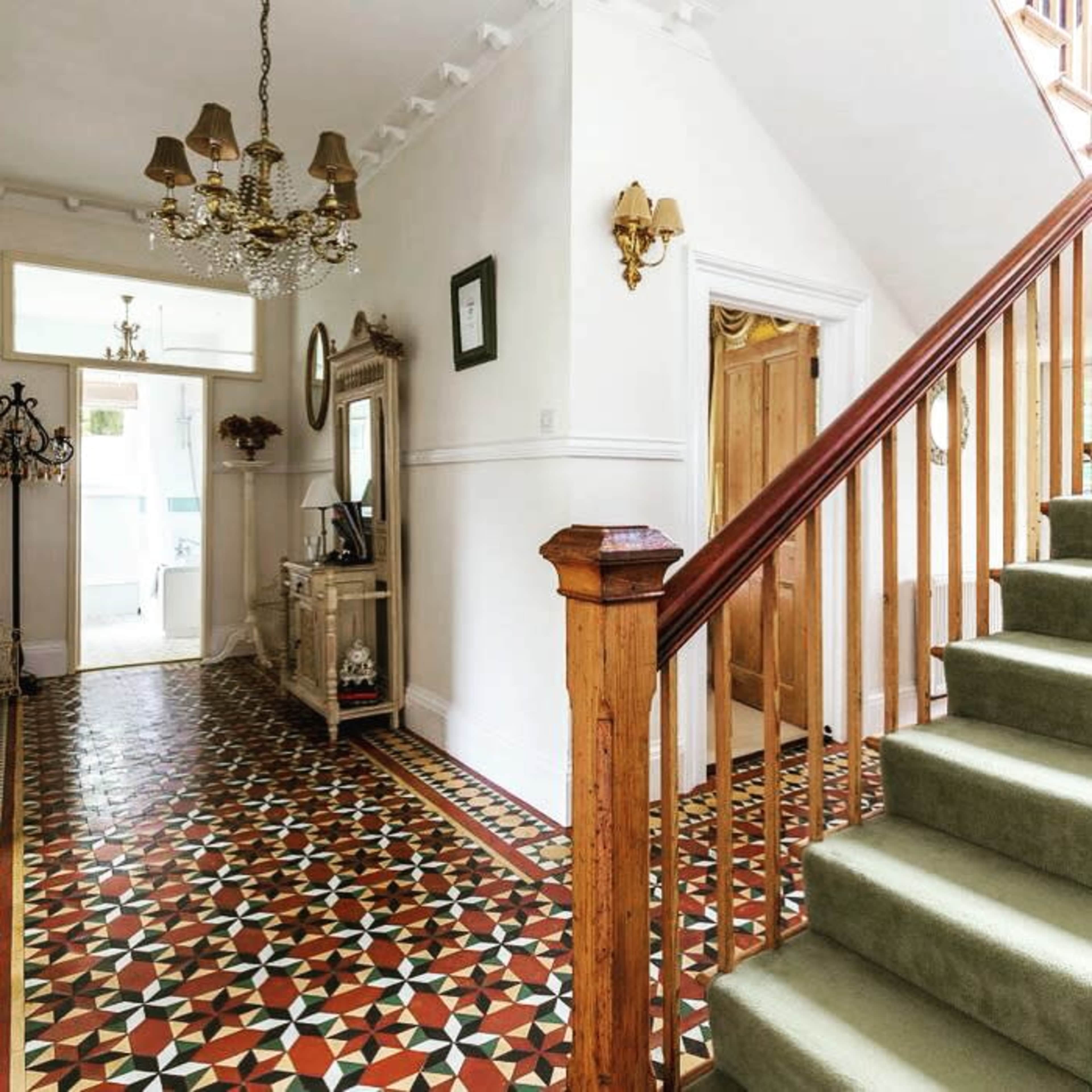 A well-lit hallway featuring a patterned tile floor, a wooden staircase, and decorative wall fixtures.