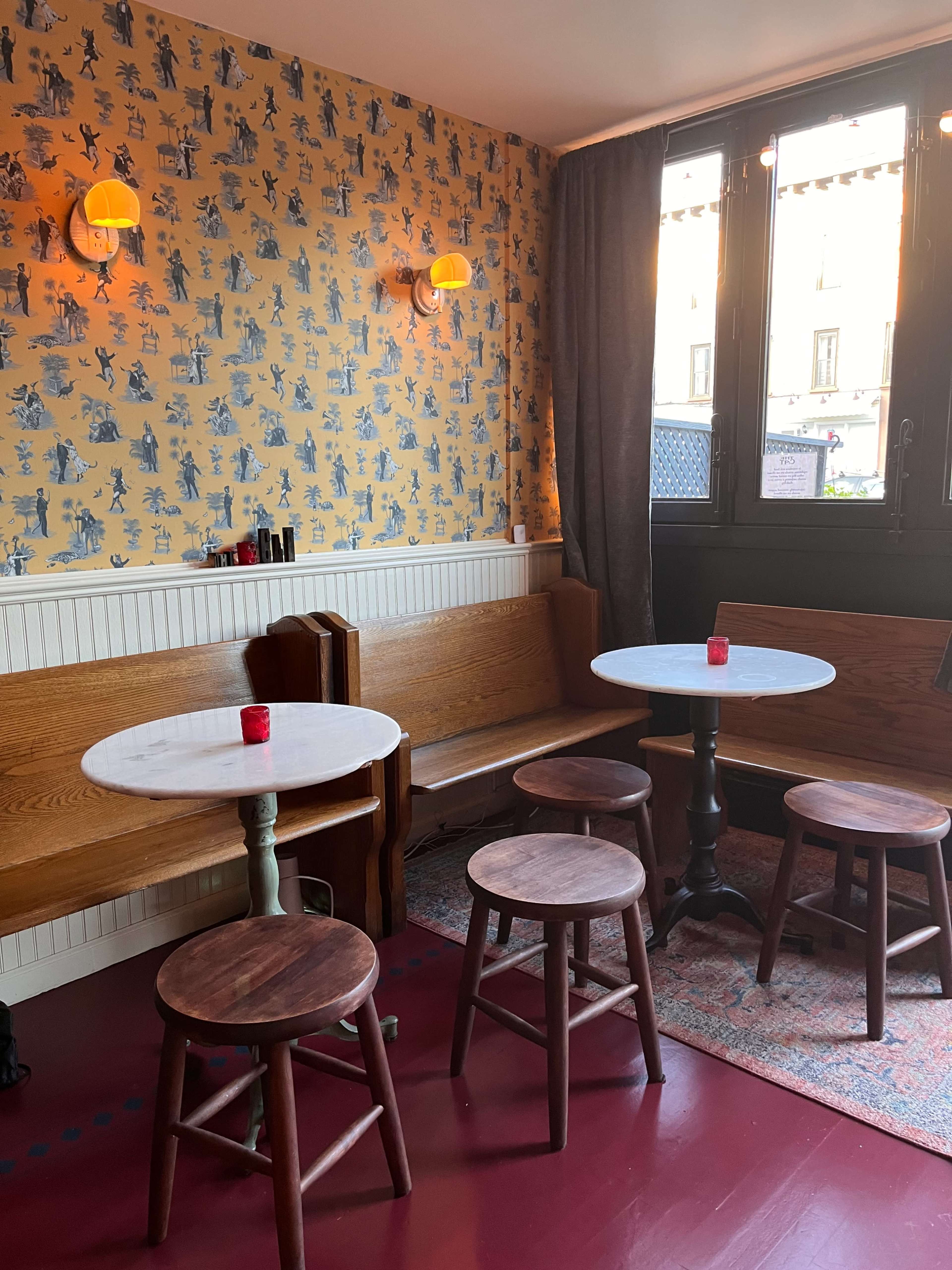 The image shows a cozy café interior with several small round tables, wooden seating along the walls, and warm lighting against a patterned wallpaper.