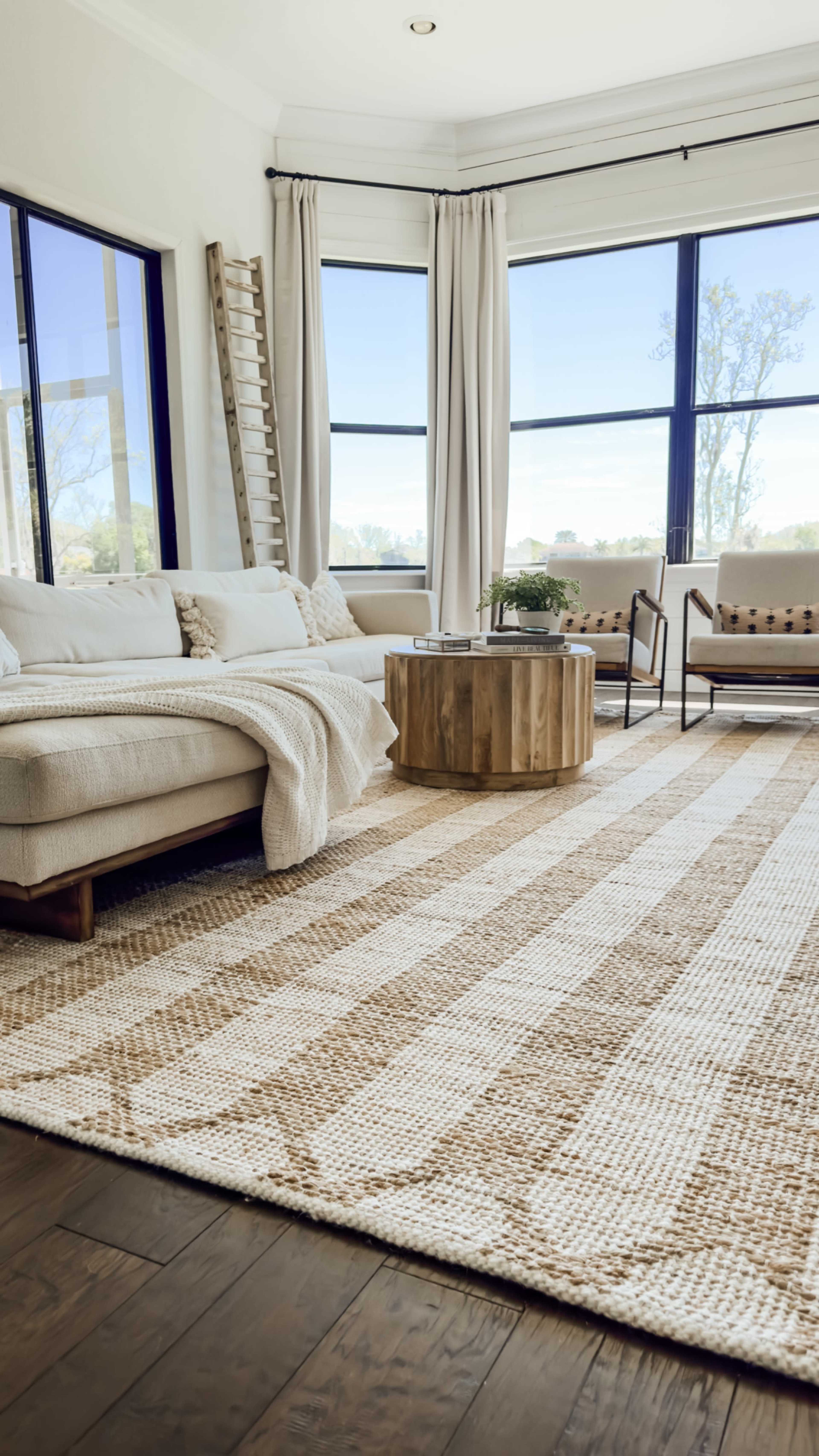 A cozy living room features a light-colored sectional sofa, a round wooden coffee table, and large windows allowing natural light to fill the space.