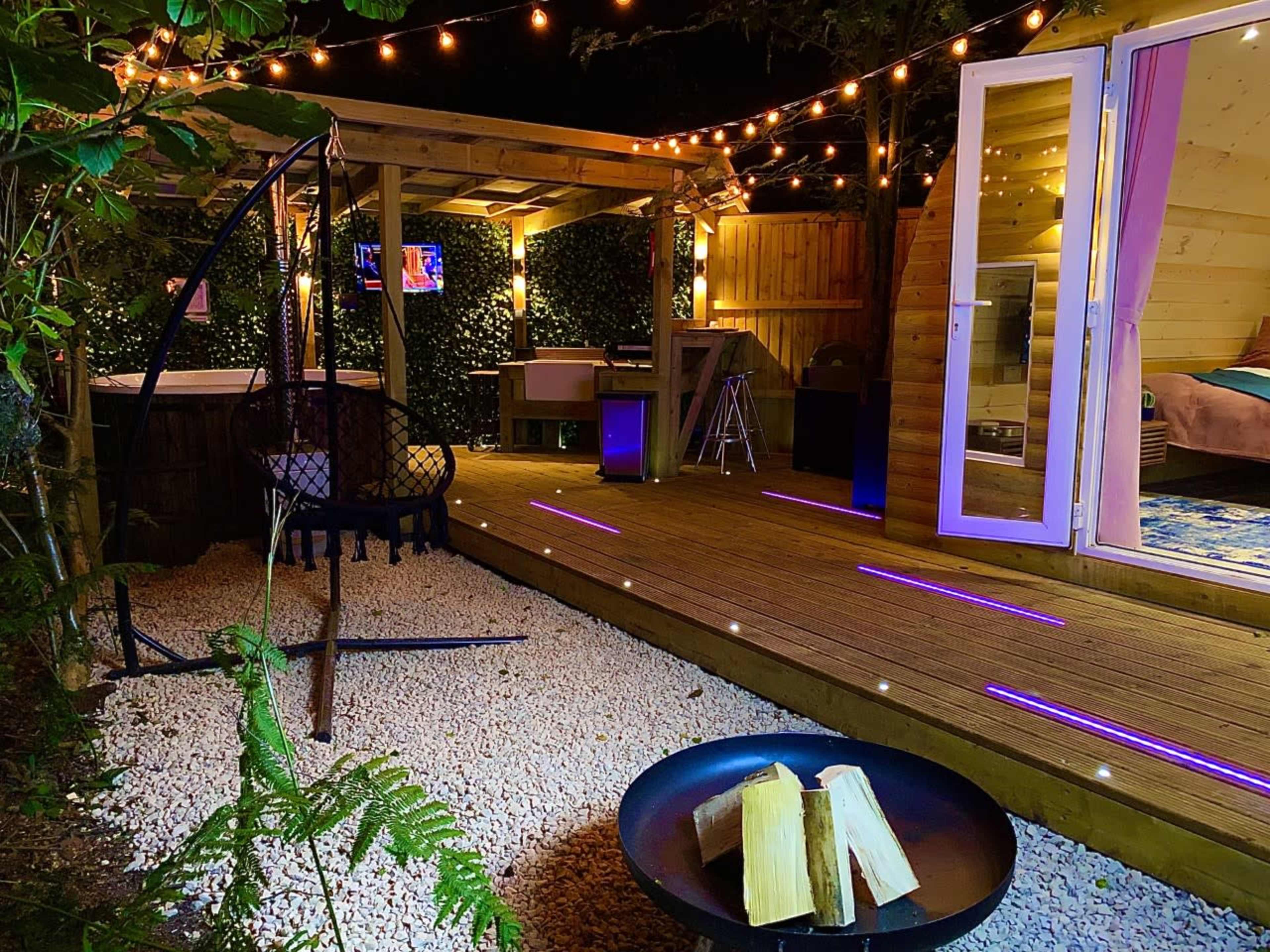A well-lit outdoor deck area featuring a hanging chair, a fire pit with logs, and a cozy cabin-style room with double doors, surrounded by greenery and string lights.