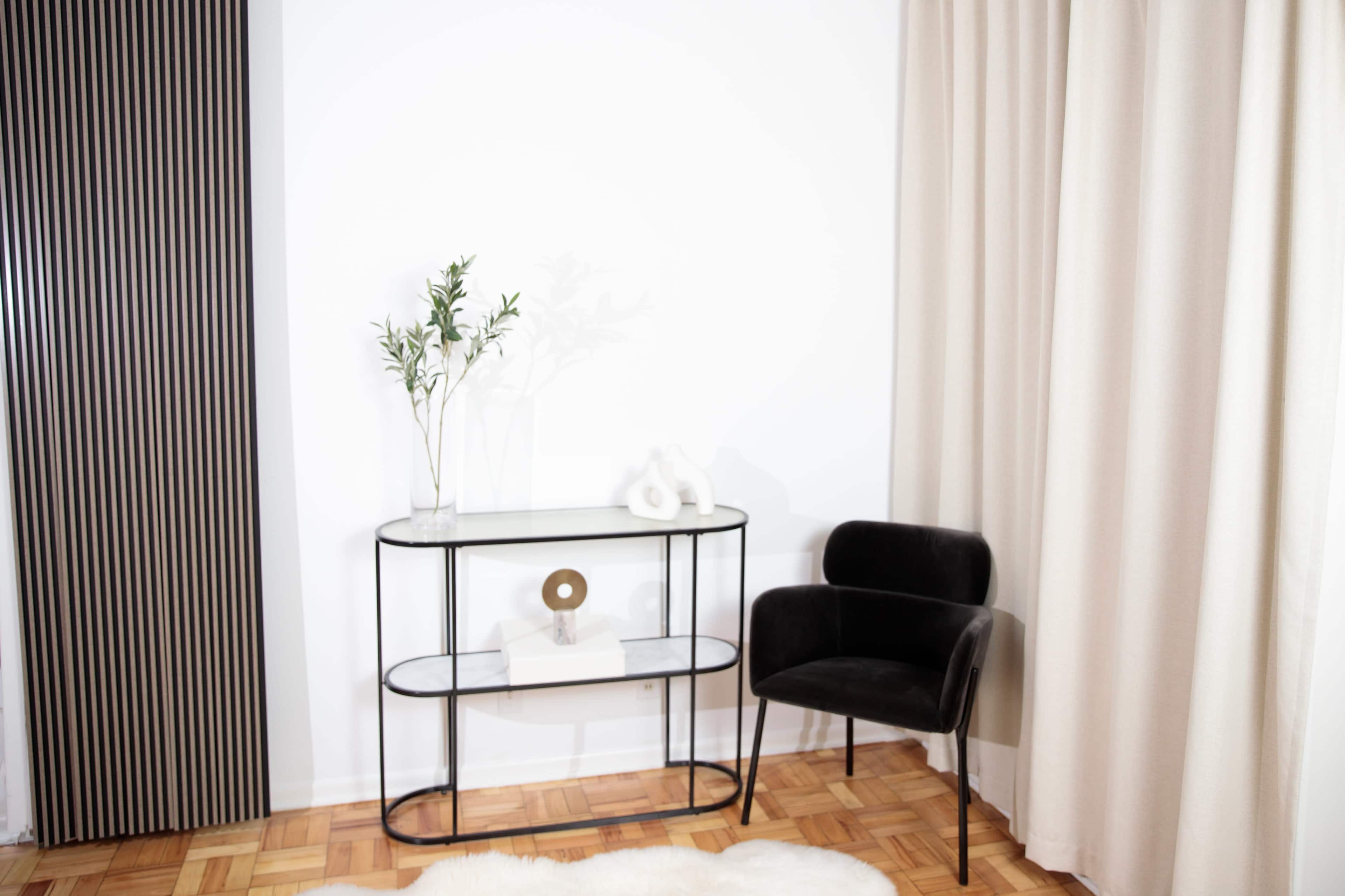 A black chair is positioned next to a glass console table against a white wall, with a decorative plant and a few accents on the table.