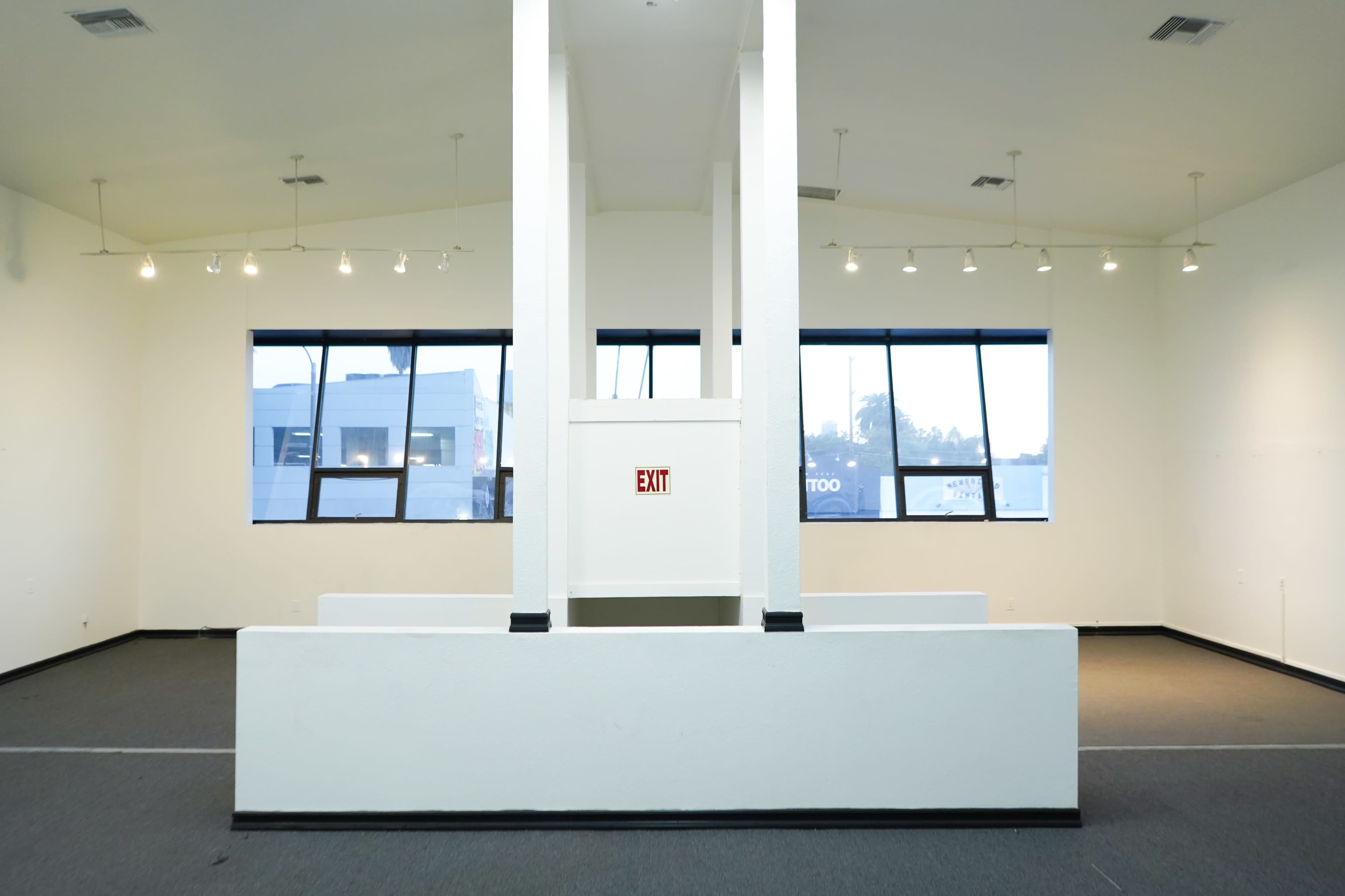 An empty room features a central white structure with an exit sign, surrounded by large windows and a muted gray carpet.