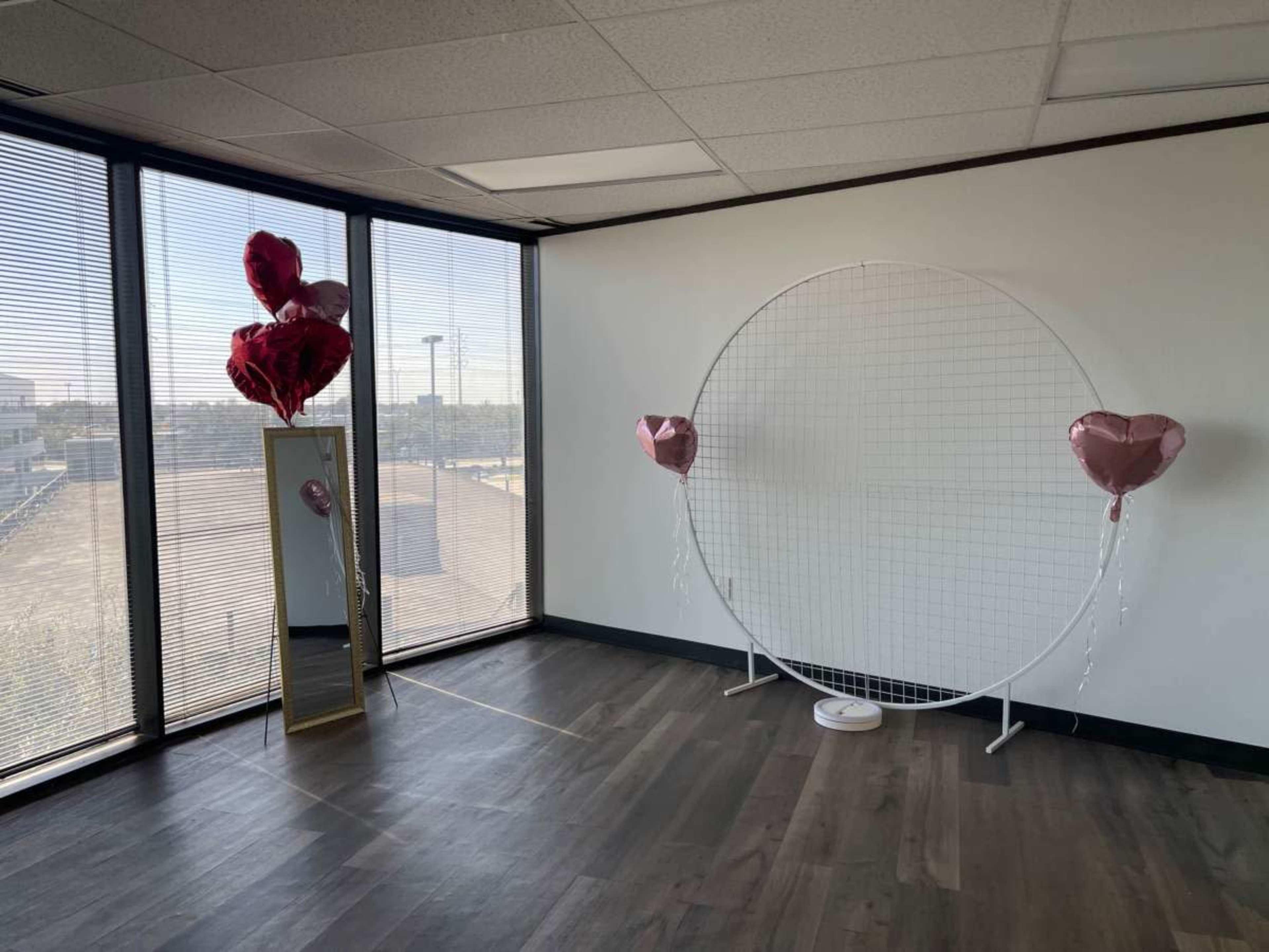 The image shows a spacious room with large windows, featuring a circular backdrop and heart-shaped balloons alongside a standing mirror.
