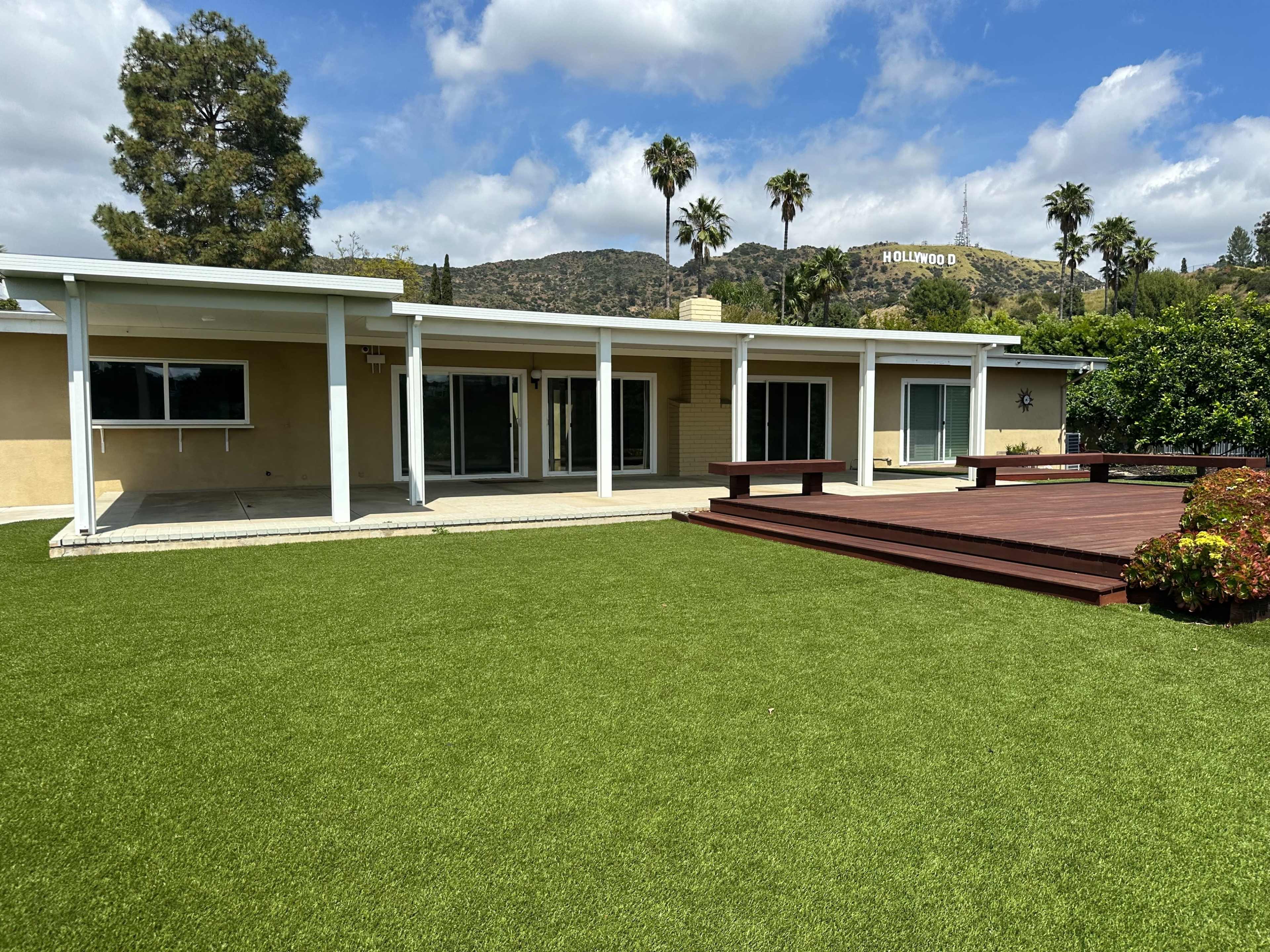A single-story house with large windows is set against a hillside displaying the Hollywood sign, surrounded by palm trees and a manicured lawn.