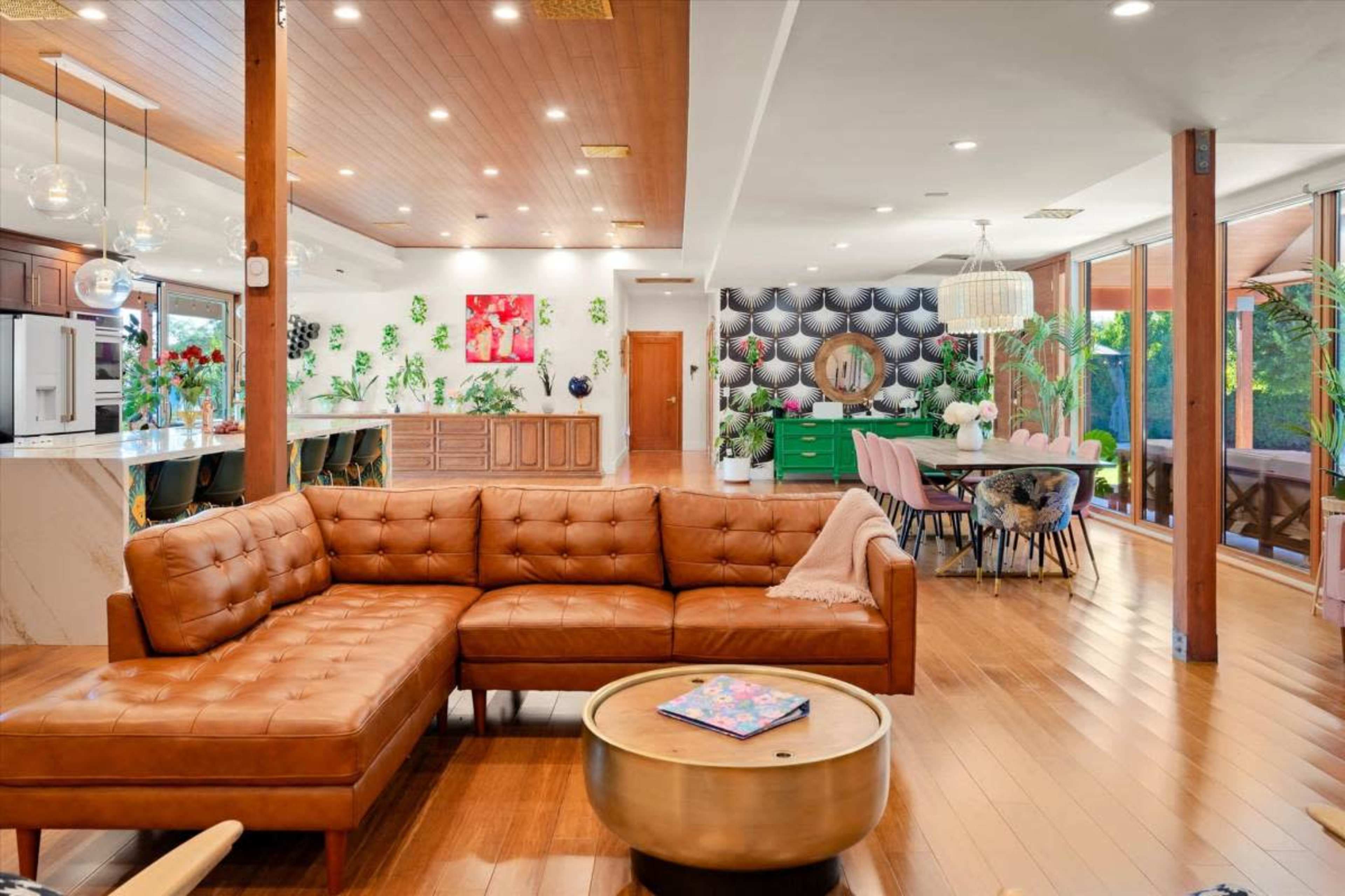 The image shows a modern, open-concept living area with a brown leather sectional sofa, a round coffee table, and a dining area featuring a green sideboard and a large wall of greenery.
