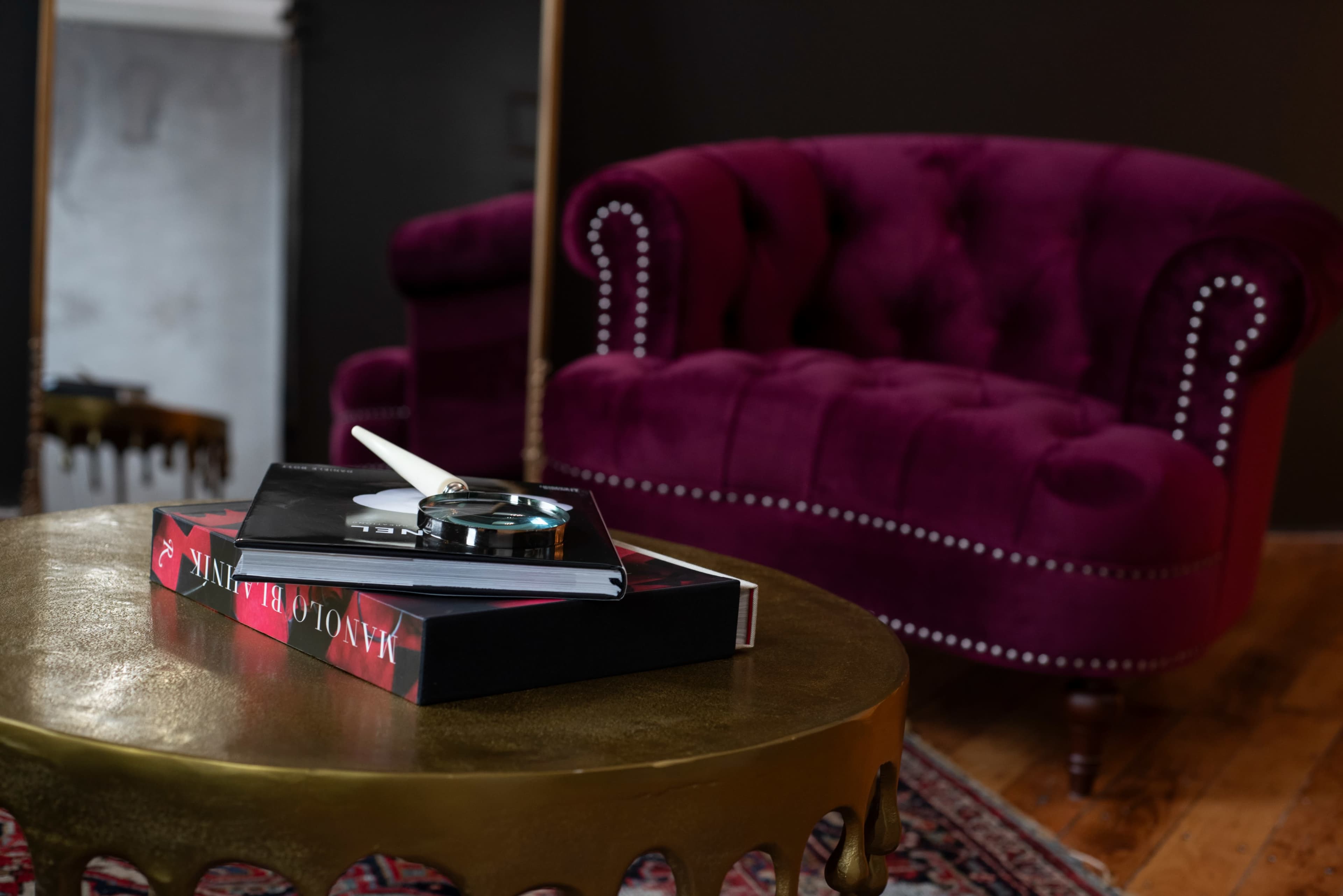 A plush purple tufted couch is set against a dark wall, with a gold coffee table displaying a book and accessories.