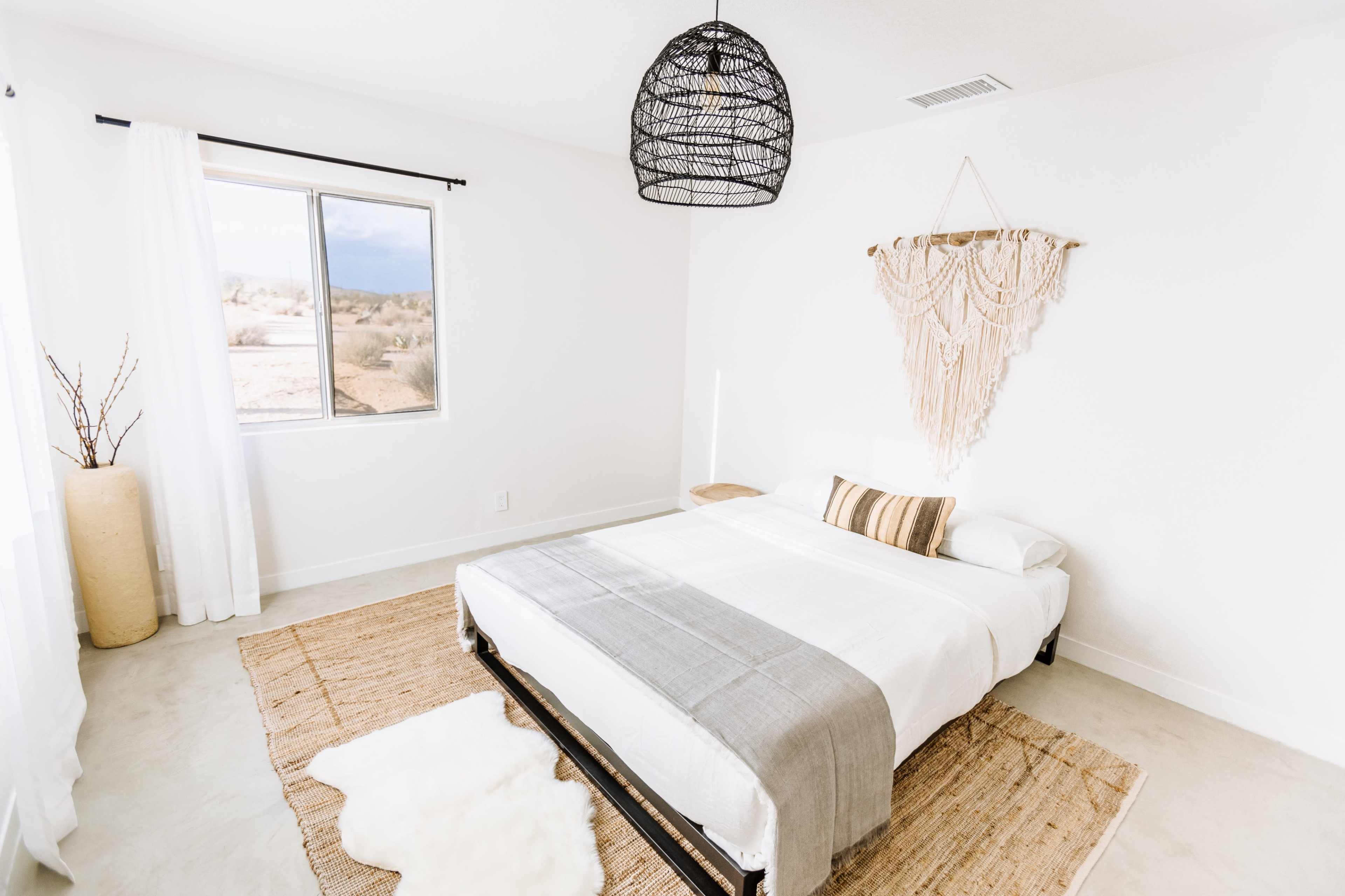 A minimalist bedroom with a bed covered in light bedding, a woven wall decoration, and a view of a landscape through a window.