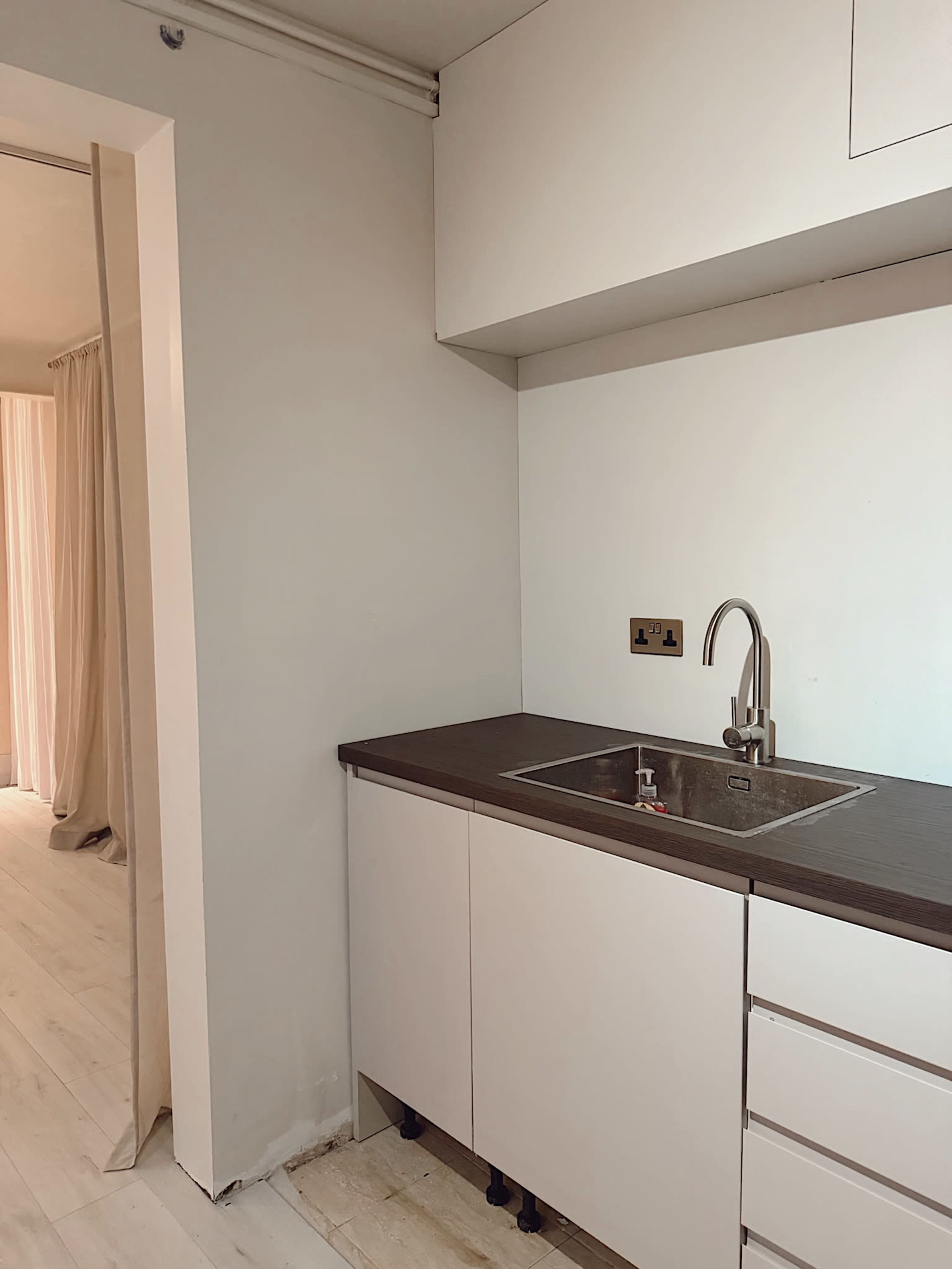 The image shows a minimalist kitchen area with a sink and cabinetry, adjacent to a doorway leading to another room.