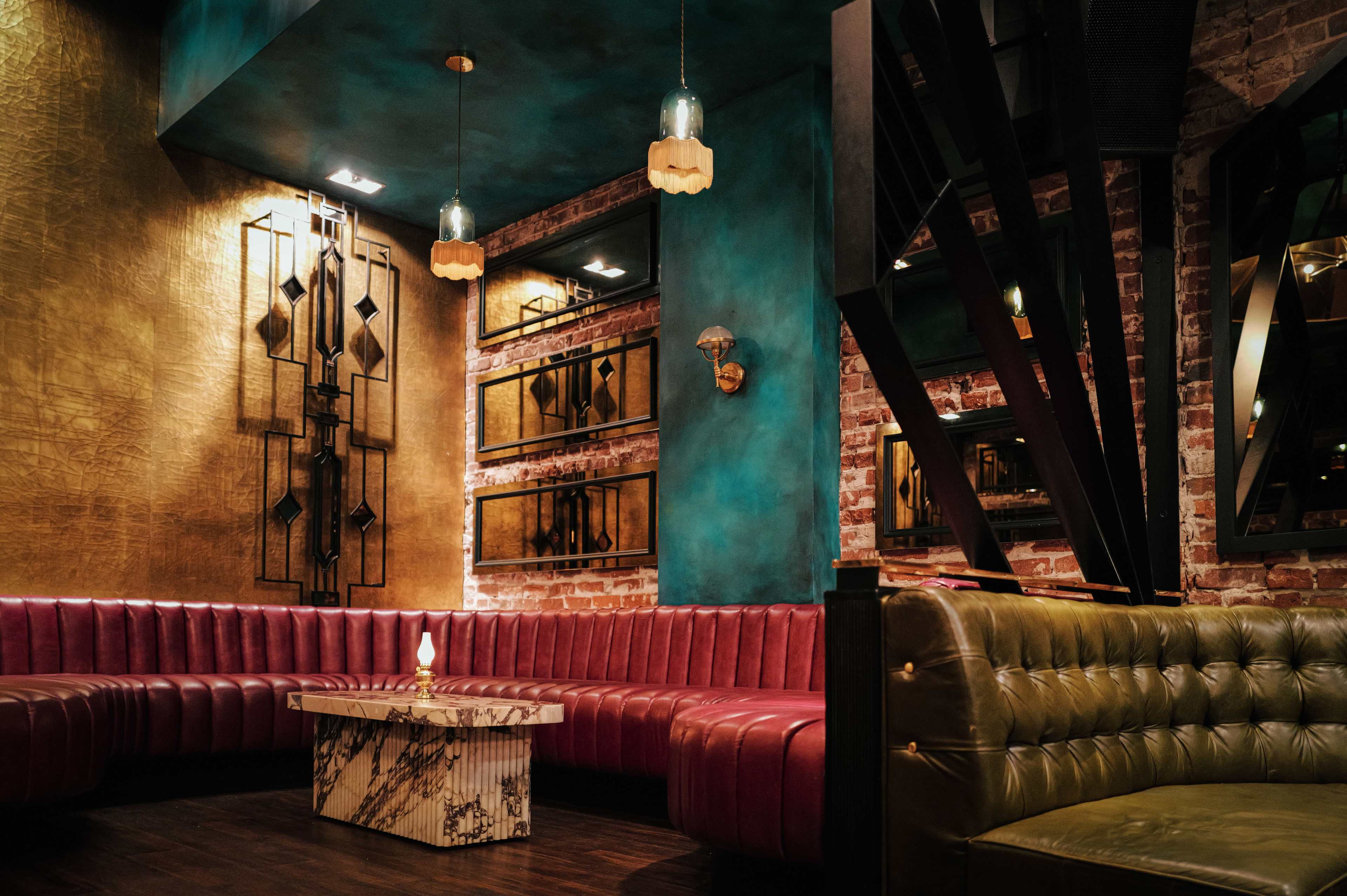 The image shows a stylish lounge with a curved pink seating area, a marble coffee table, and decorative lighting against a backdrop of exposed brick and vibrant wall colors.