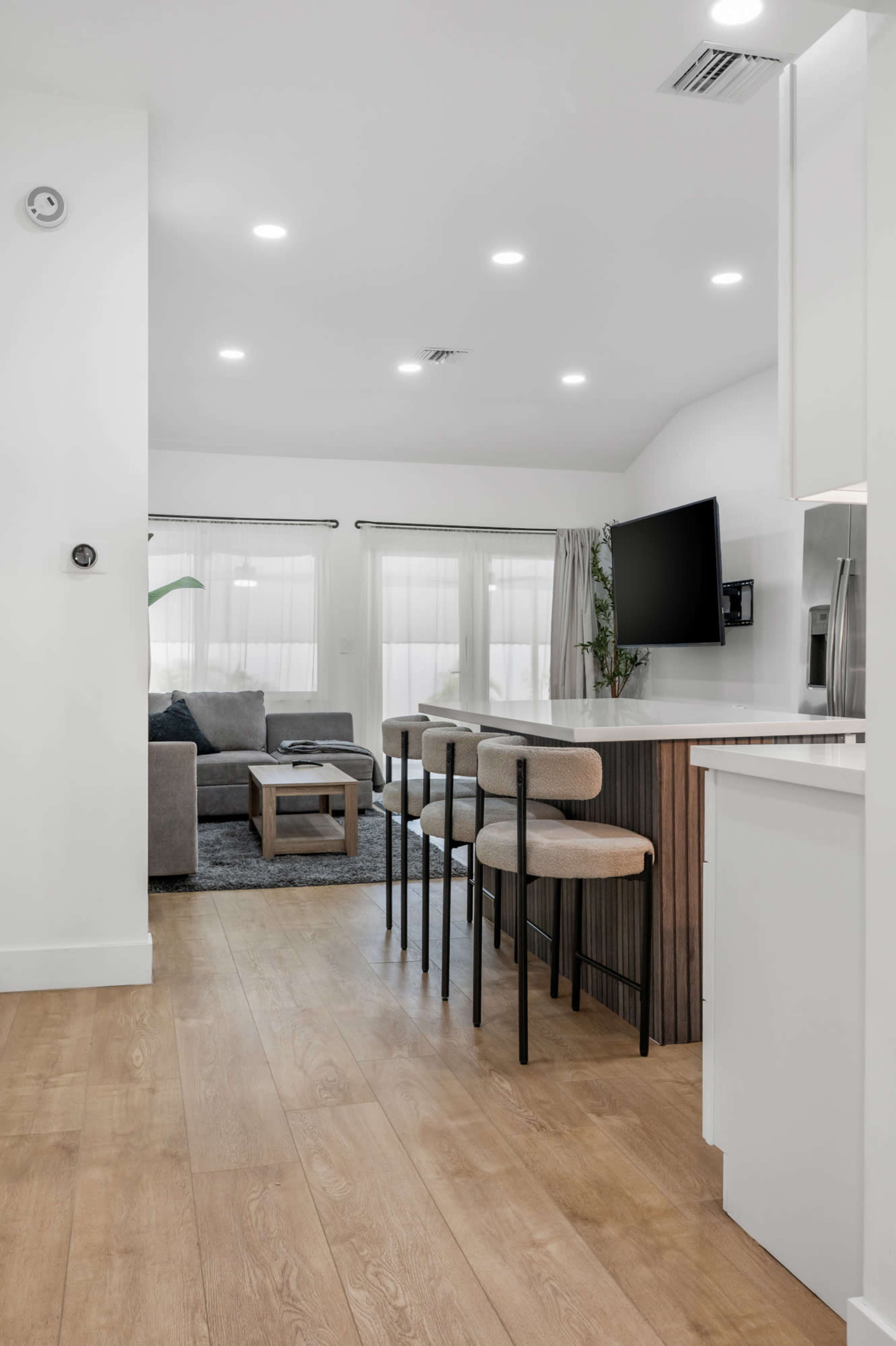 A modern kitchen and living area features a sleek countertop with stools, a gray sofa, and large windows allowing natural light.