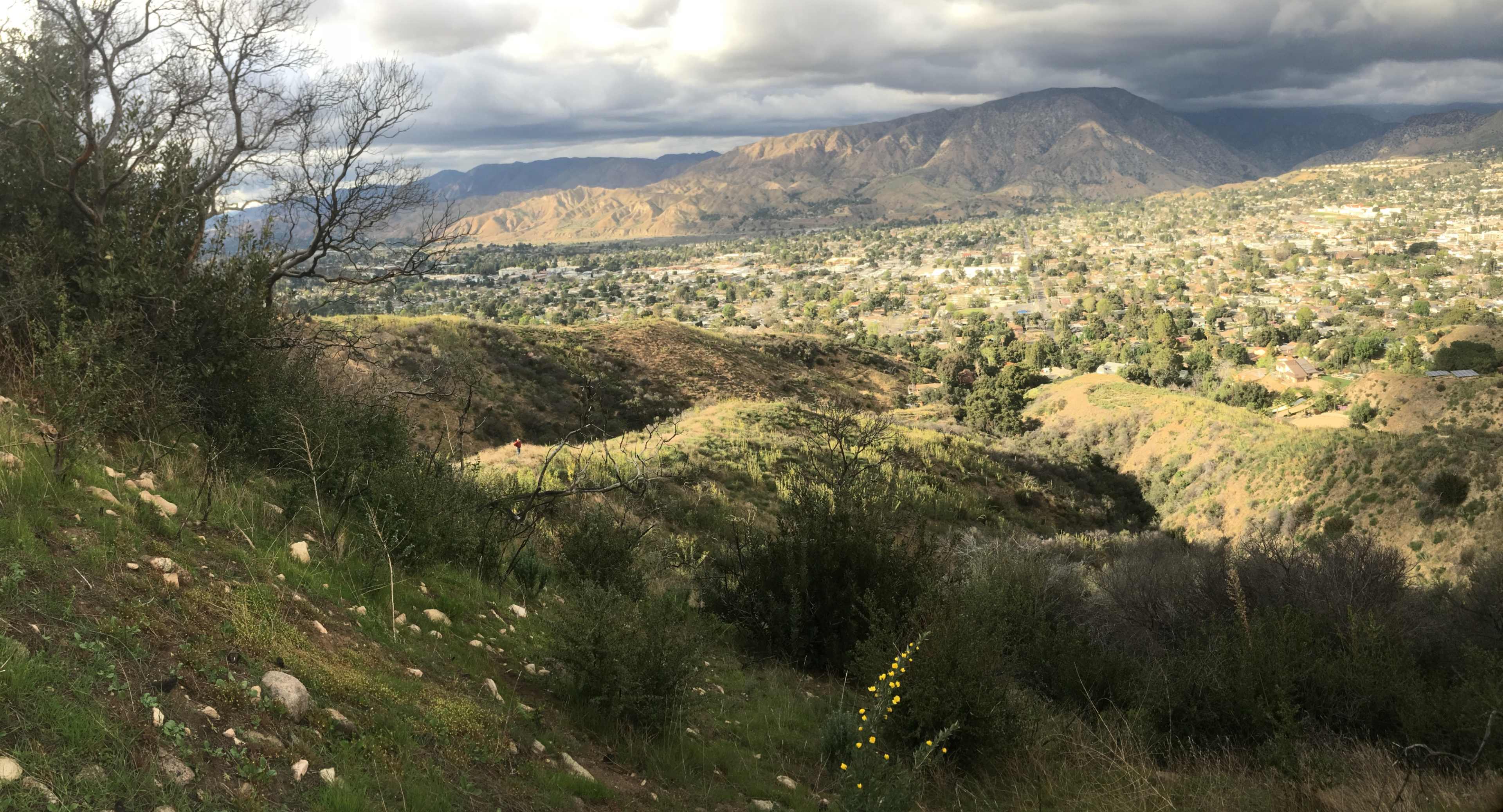 A hilly landscape with a view of a valley and mountains in the background, under a cloudy sky.
