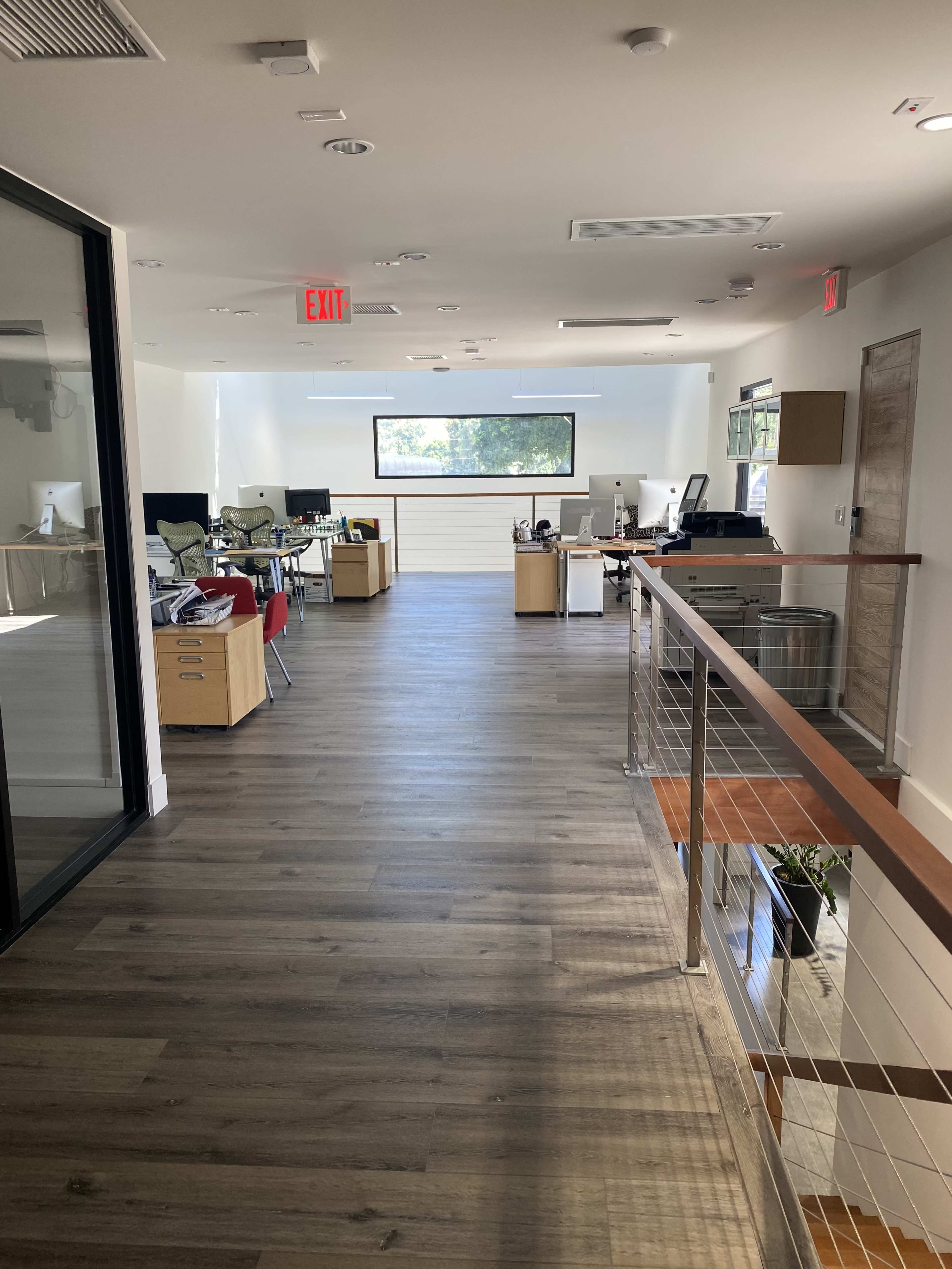 A modern office interior with desks, chairs, and a large window along a wooden floor corridor.