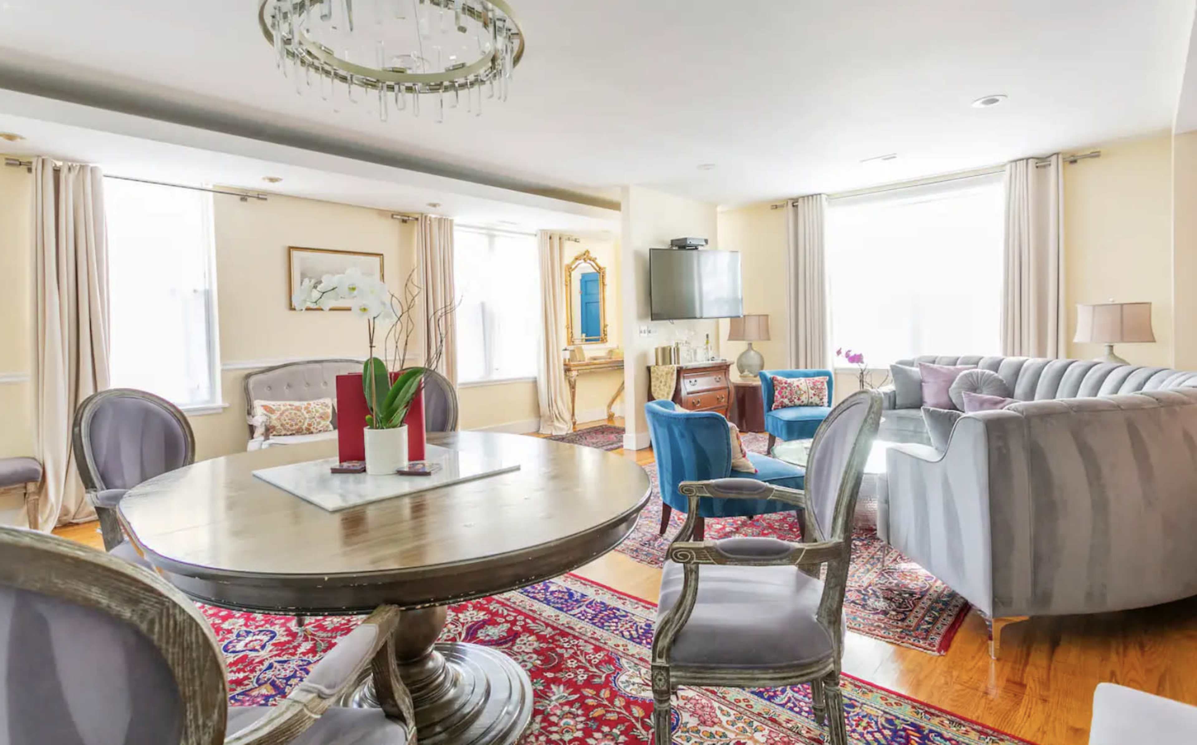 A living room features a round dining table, a gray sectional sofa, and large windows illuminating the space.