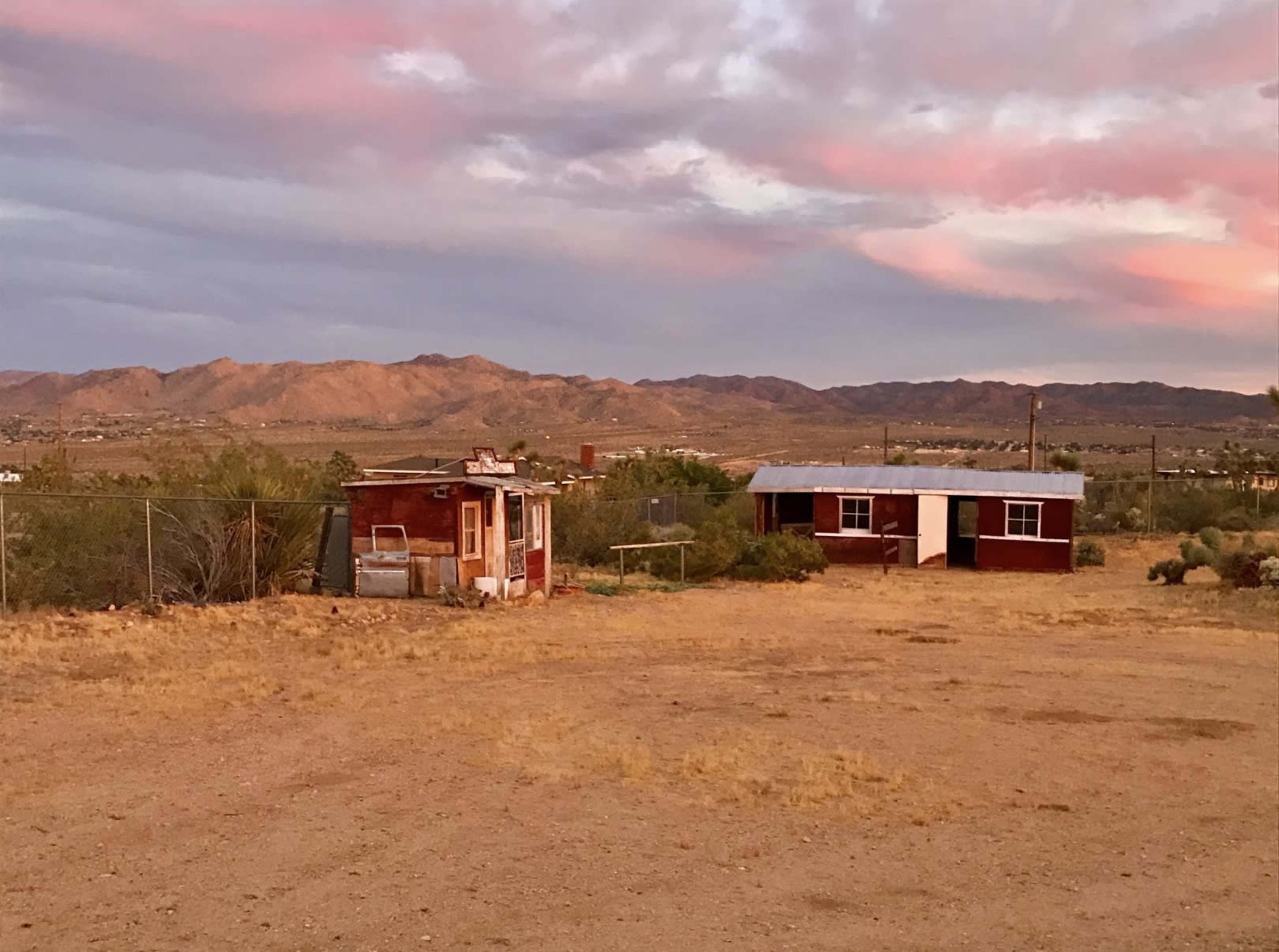 A weathered red cabin and a small structure are set against a backdrop of distant mountains under a cloudy sky.