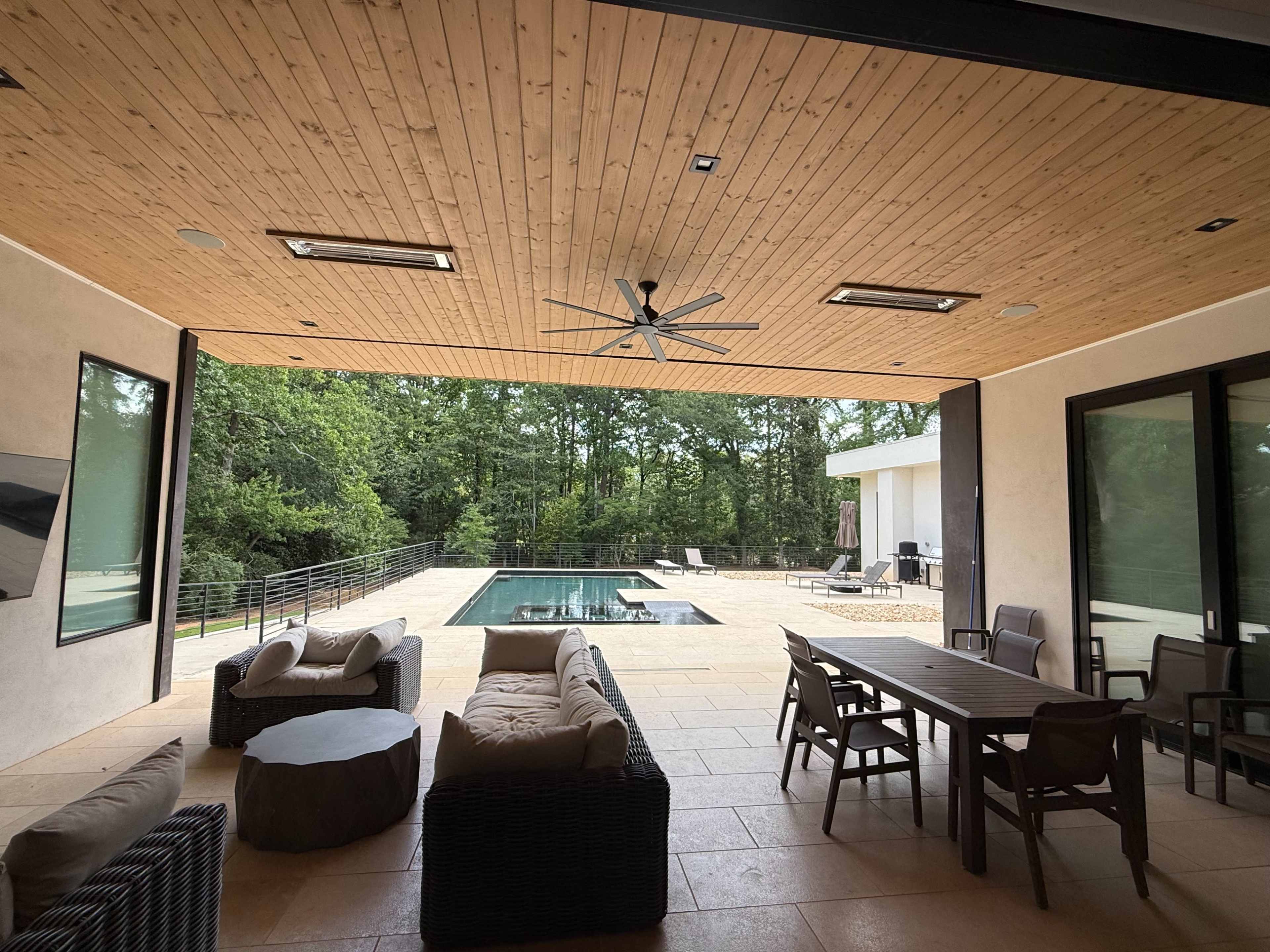 The image shows a modern outdoor patio area with a wooden ceiling, seating, and a pool surrounded by trees and lounge chairs.