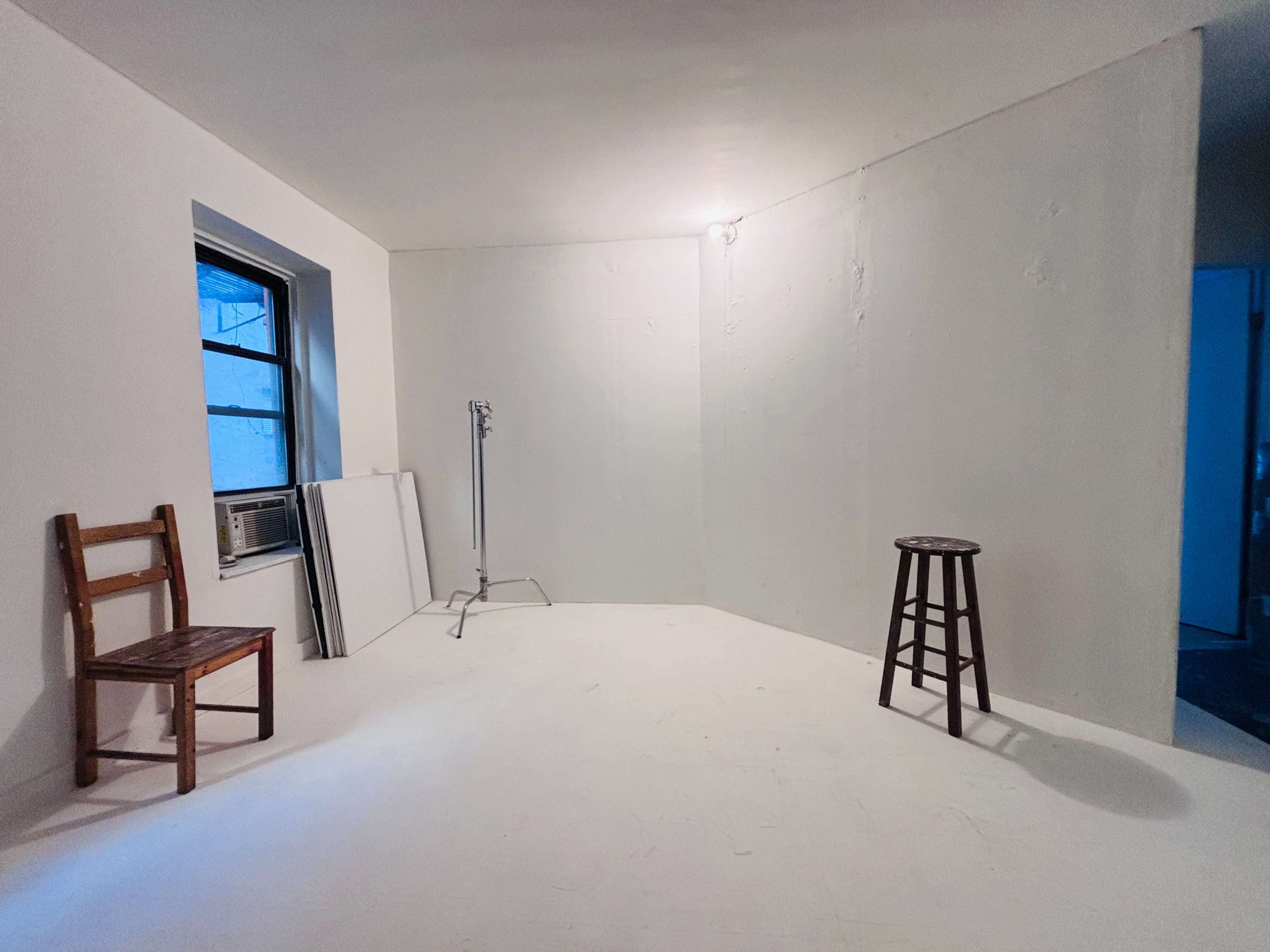 The image shows a sparsely furnished room with a wooden chair, a stool, an air conditioning unit, and a light stand against plain white walls.