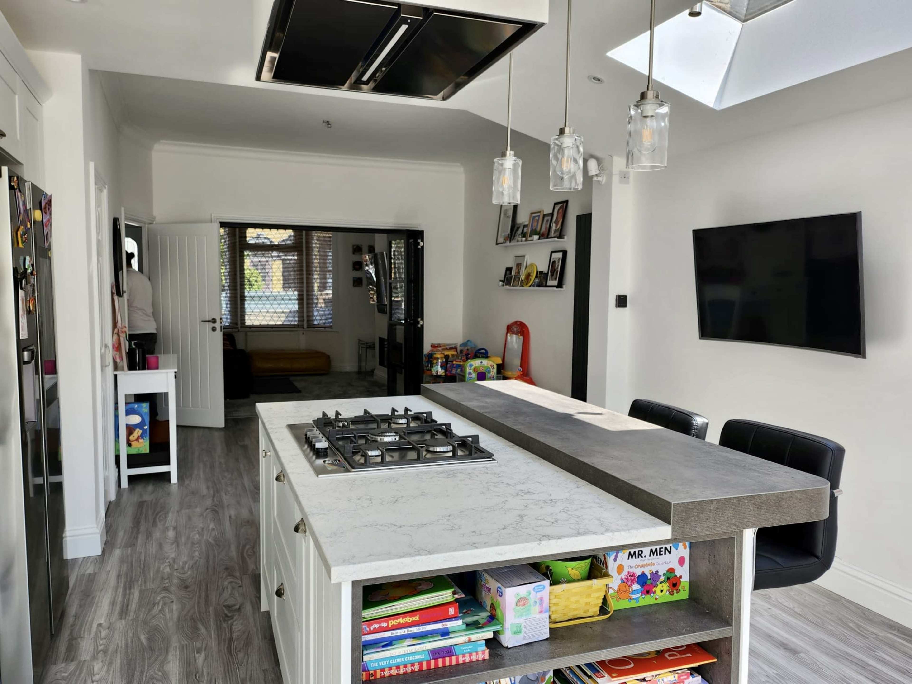 The image shows a modern kitchen with a central island featuring a gas stove, surrounded by books and toys, and an adjoining living area visible in the background.