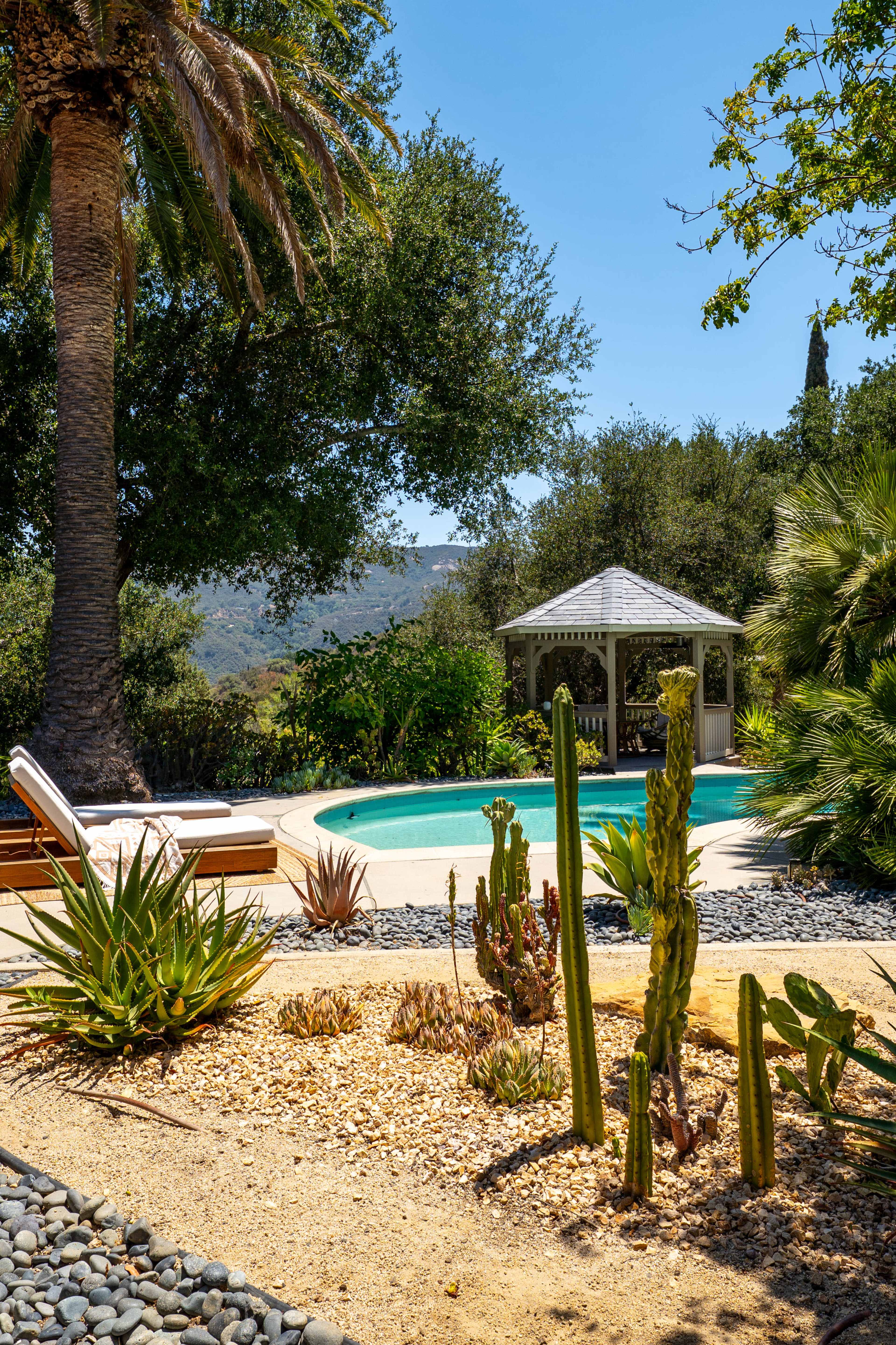 The scene features a swimming pool surrounded by various desert plants and a gazebo, set against a backdrop of trees and distant mountains under a clear blue sky.