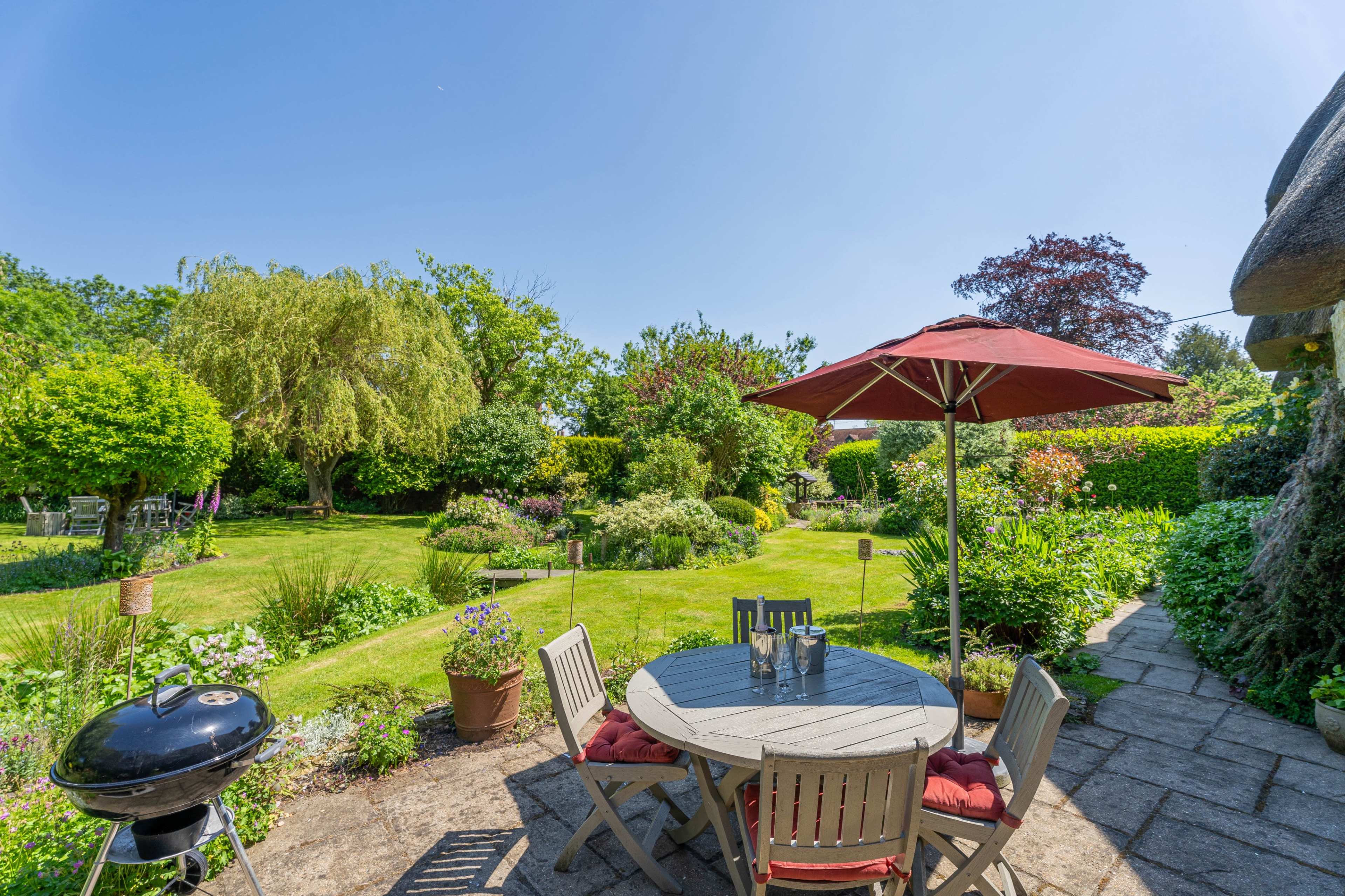 A patio with a table and an umbrella overlooks a well-maintained garden featuring various trees and plants.