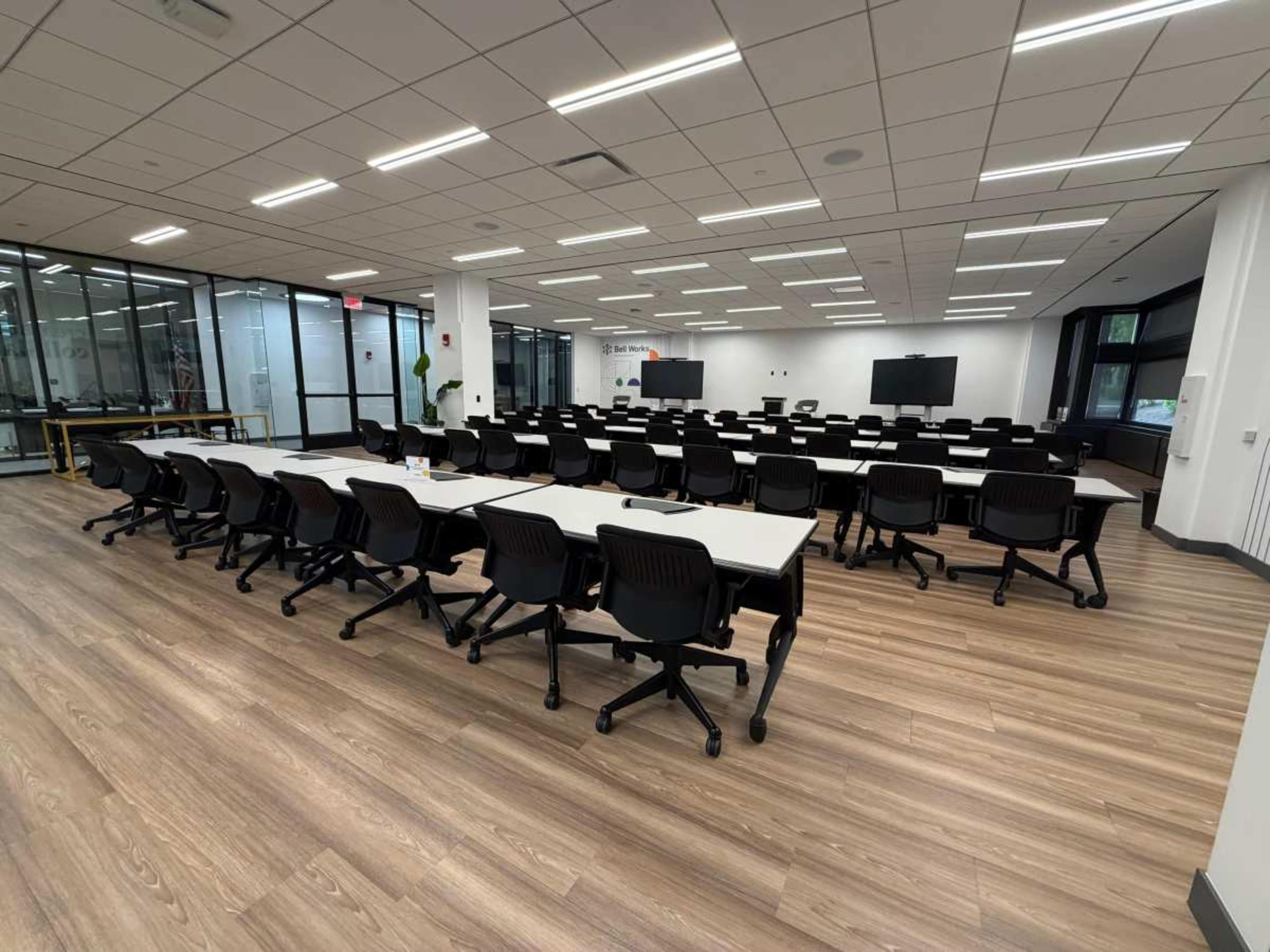 The image shows a large, empty classroom with multiple rows of tables and chairs arranged for a workshop or seminar, illuminated by bright overhead lighting.