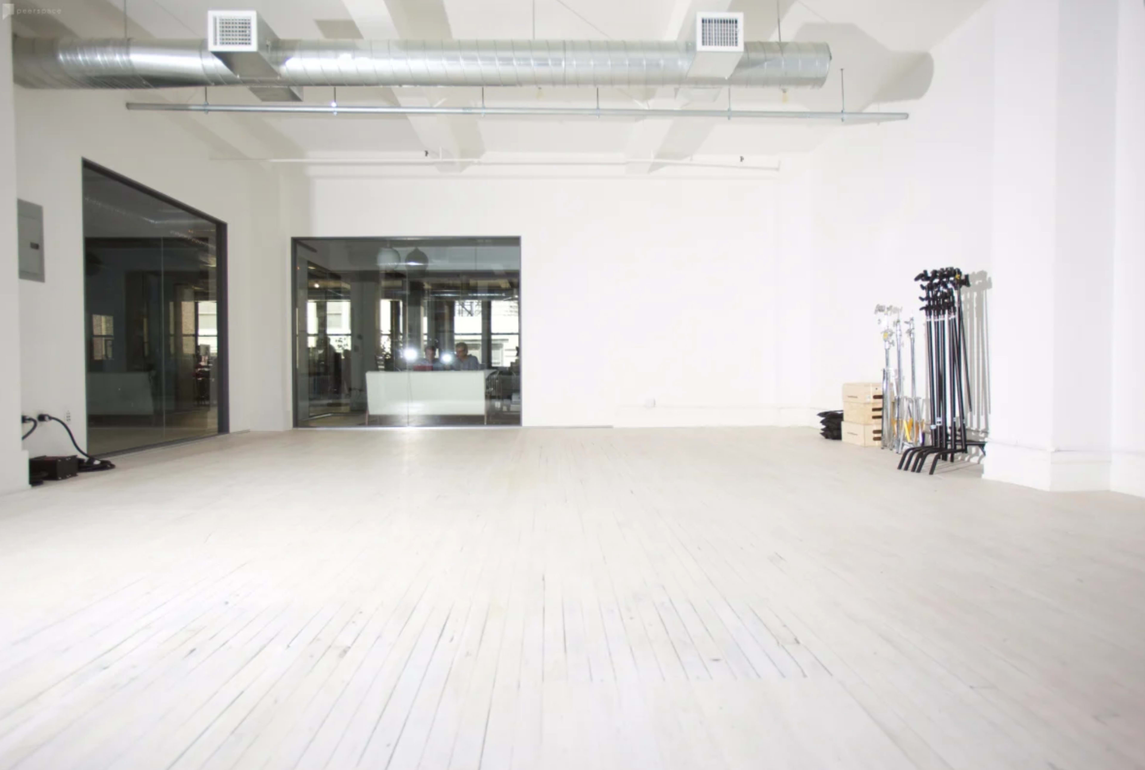 A spacious and empty room with light wood flooring and large windows, featuring a conference area visible through glass walls and equipment neatly arranged in one corner.