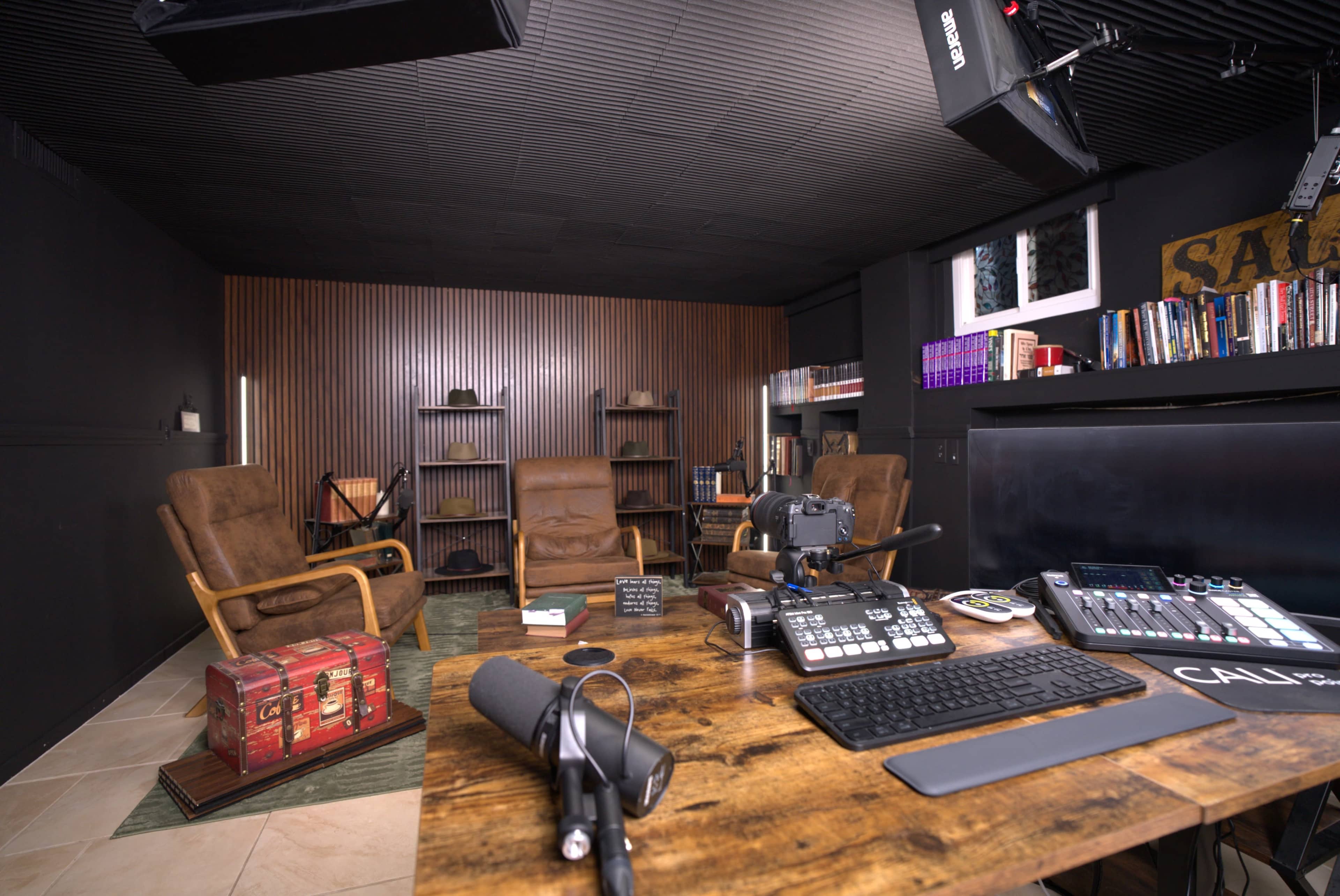 The image shows a recording studio with three armchairs, bookshelves, a wooden desk equipped with audio equipment, and a vintage trunk.