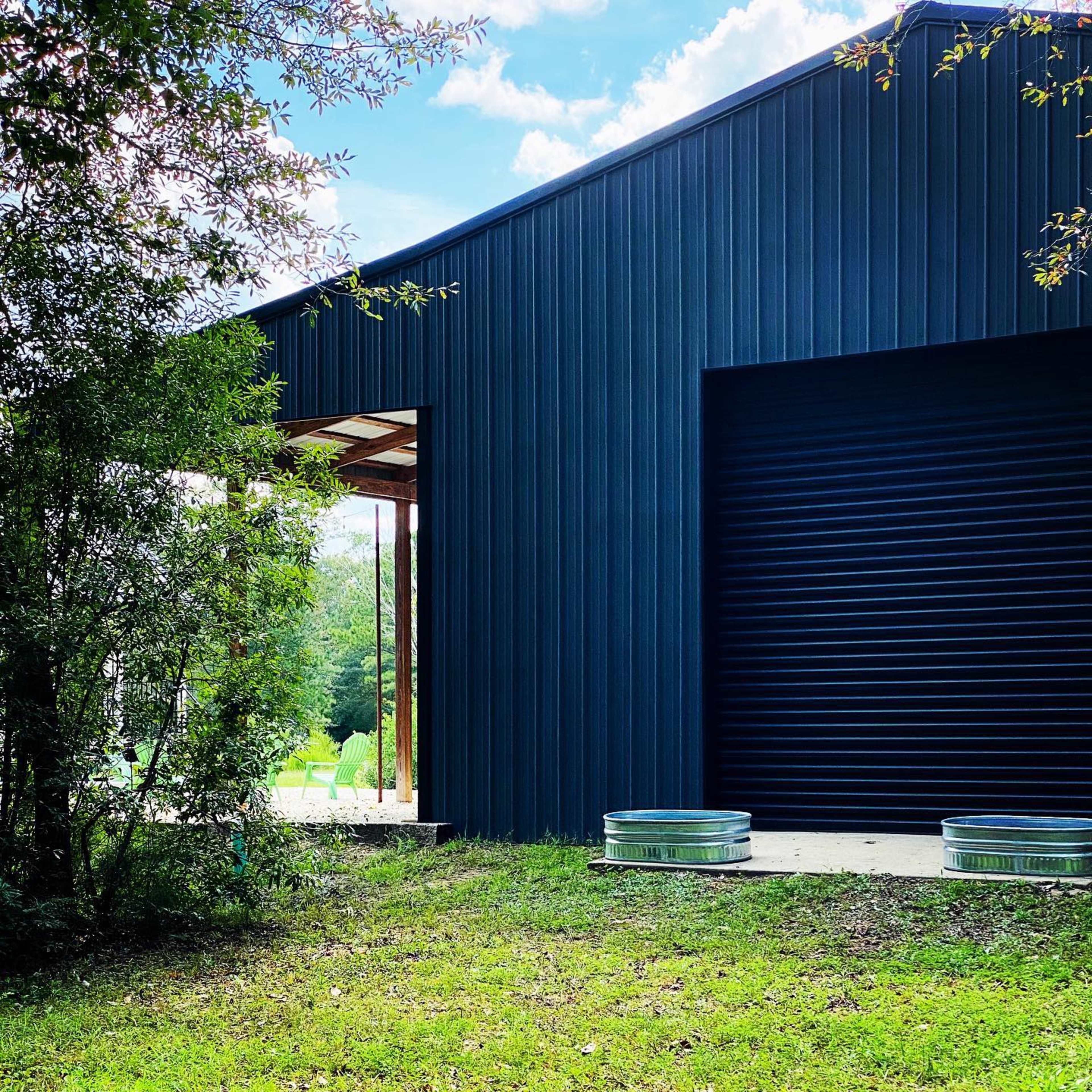 A dark metal building features a large roll-up door and is surrounded by greenery and grass.