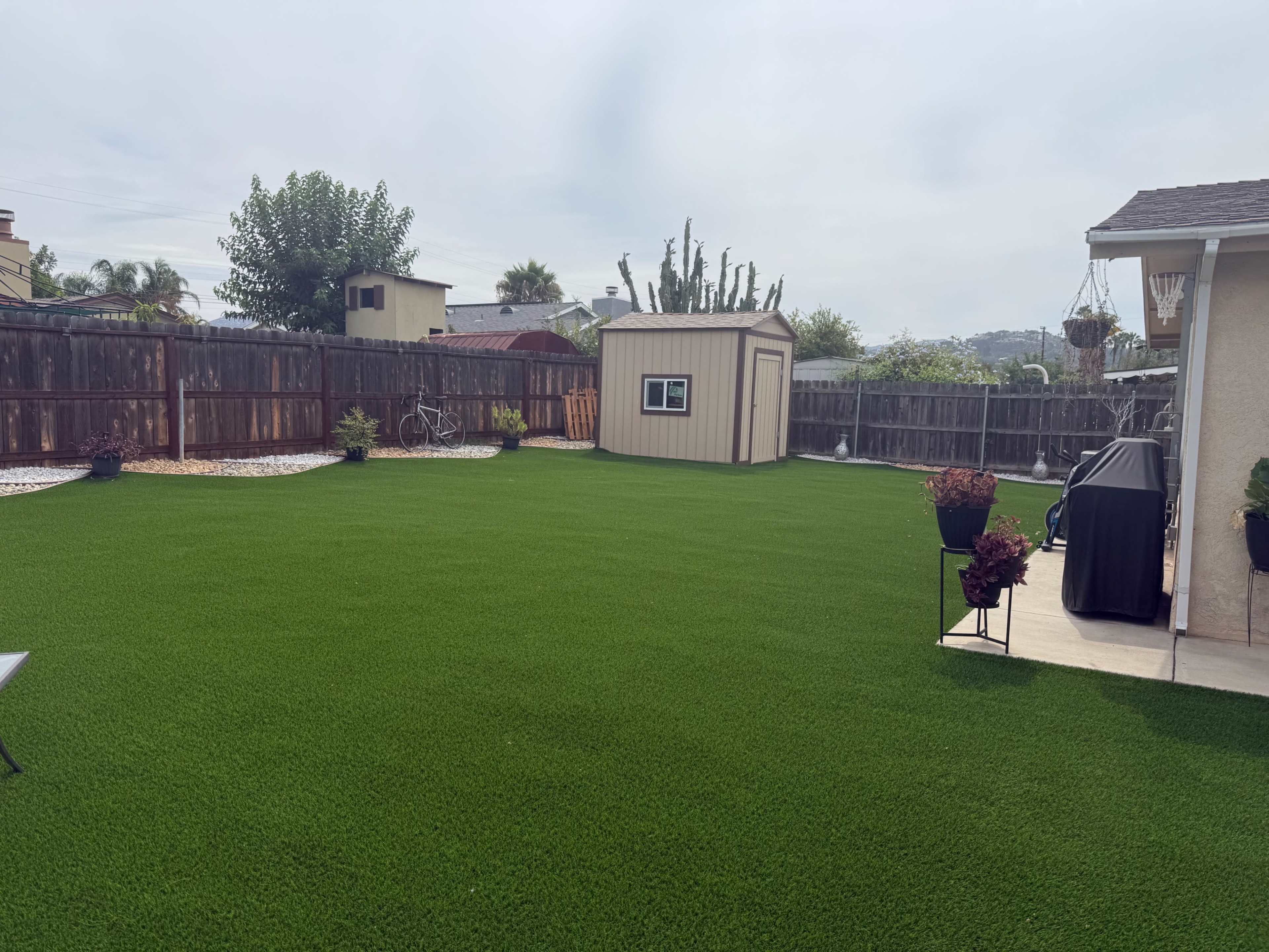 The image depicts a backyard featuring a neatly maintained artificial lawn, a small shed, and a patio area with planters and a grill.