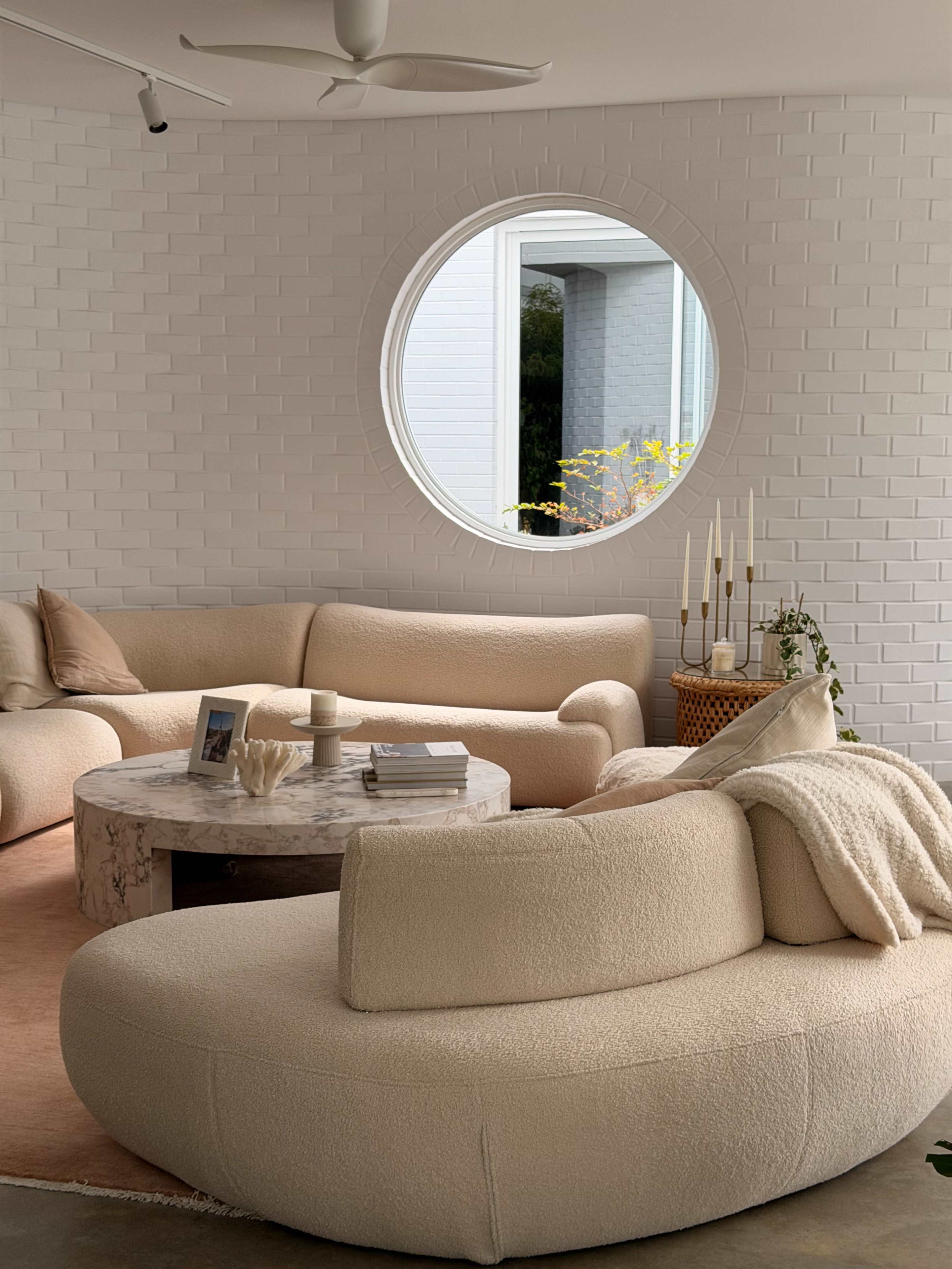 A living room features curved, light-colored sofas, a circular marble coffee table, and a large round window that lets in natural light.