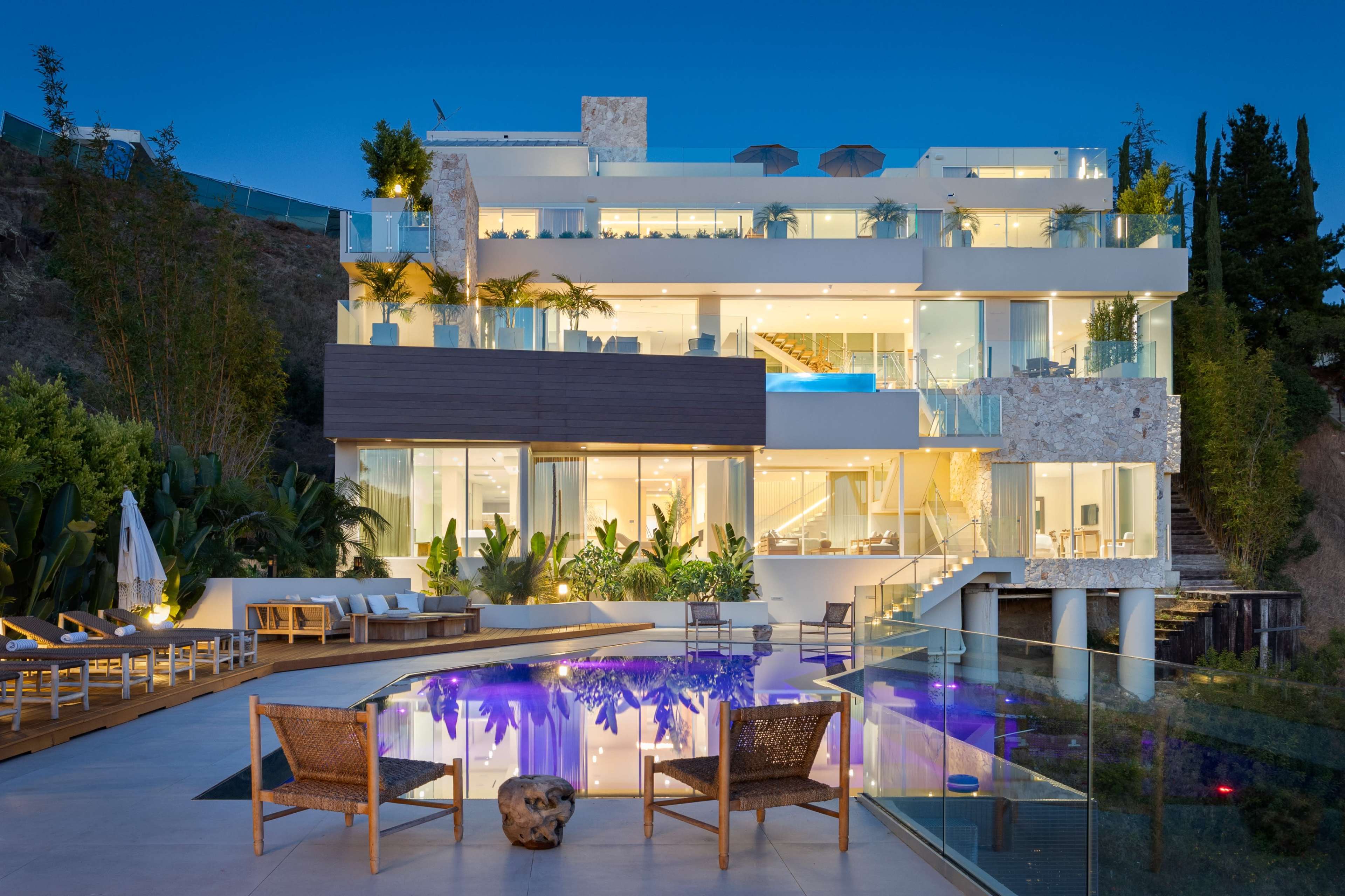 The image shows a modern three-story house with large glass windows, surrounded by greenery and featuring an outdoor pool.