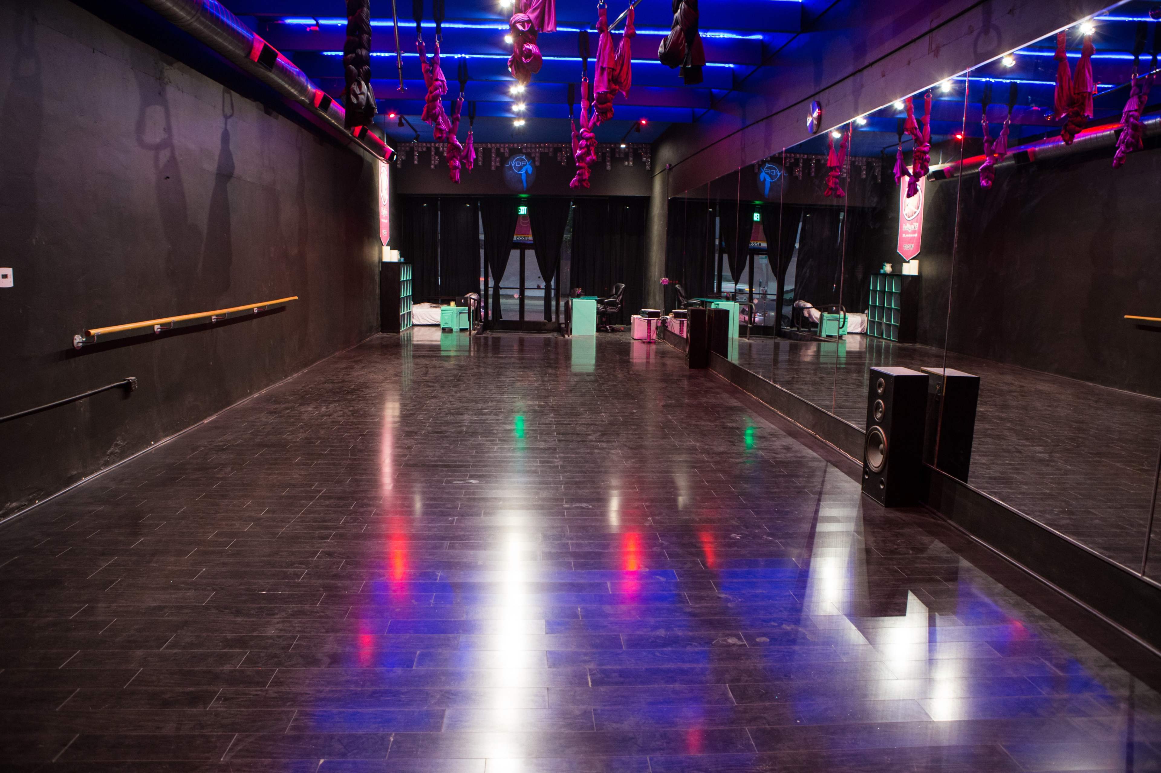 A spacious dance studio with a mirrored wall, hardwood flooring, and colorful lighting overhead.