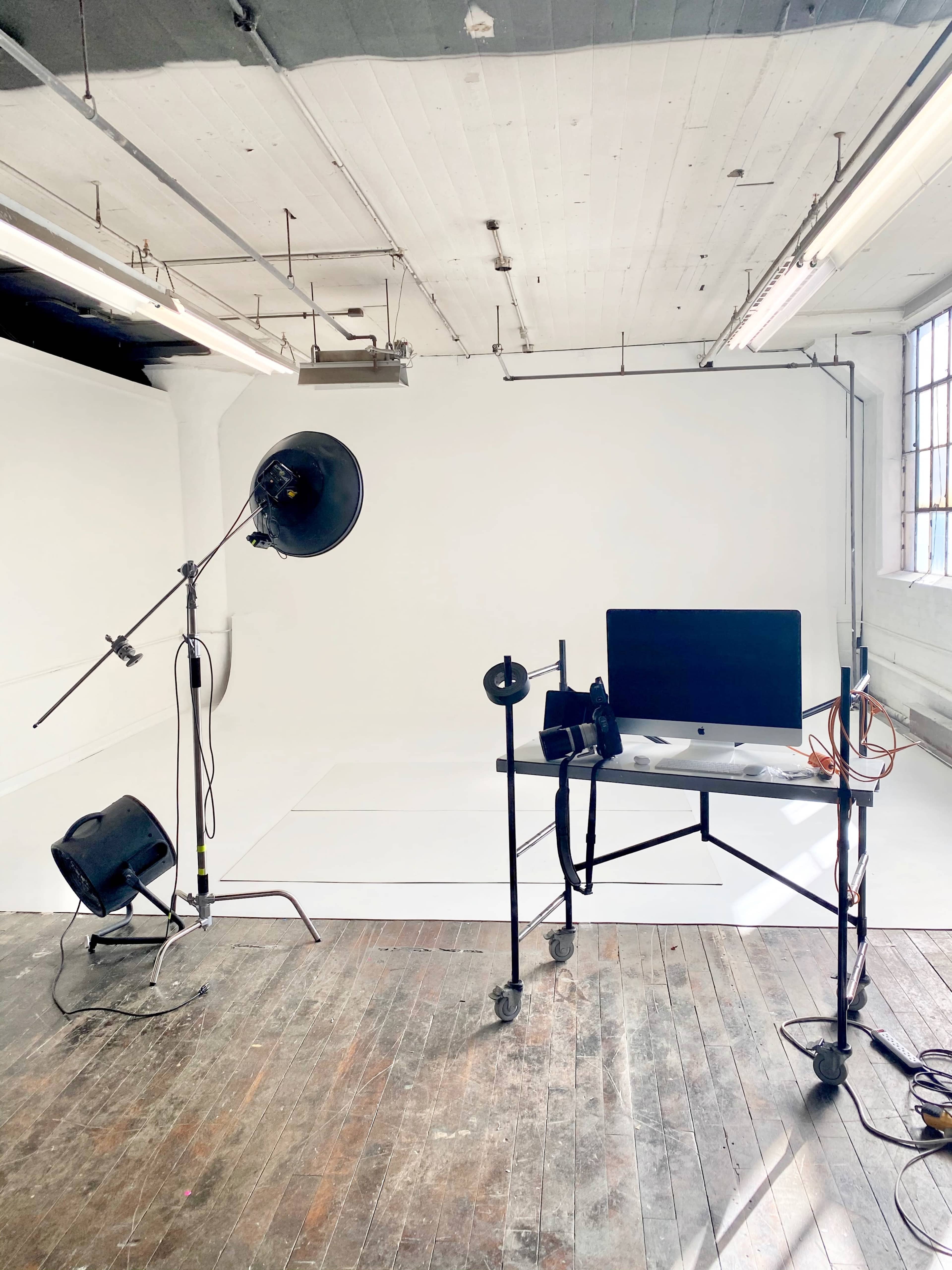 A photography studio with a blank white backdrop, a light stand, and a desk featuring a computer on wheels.