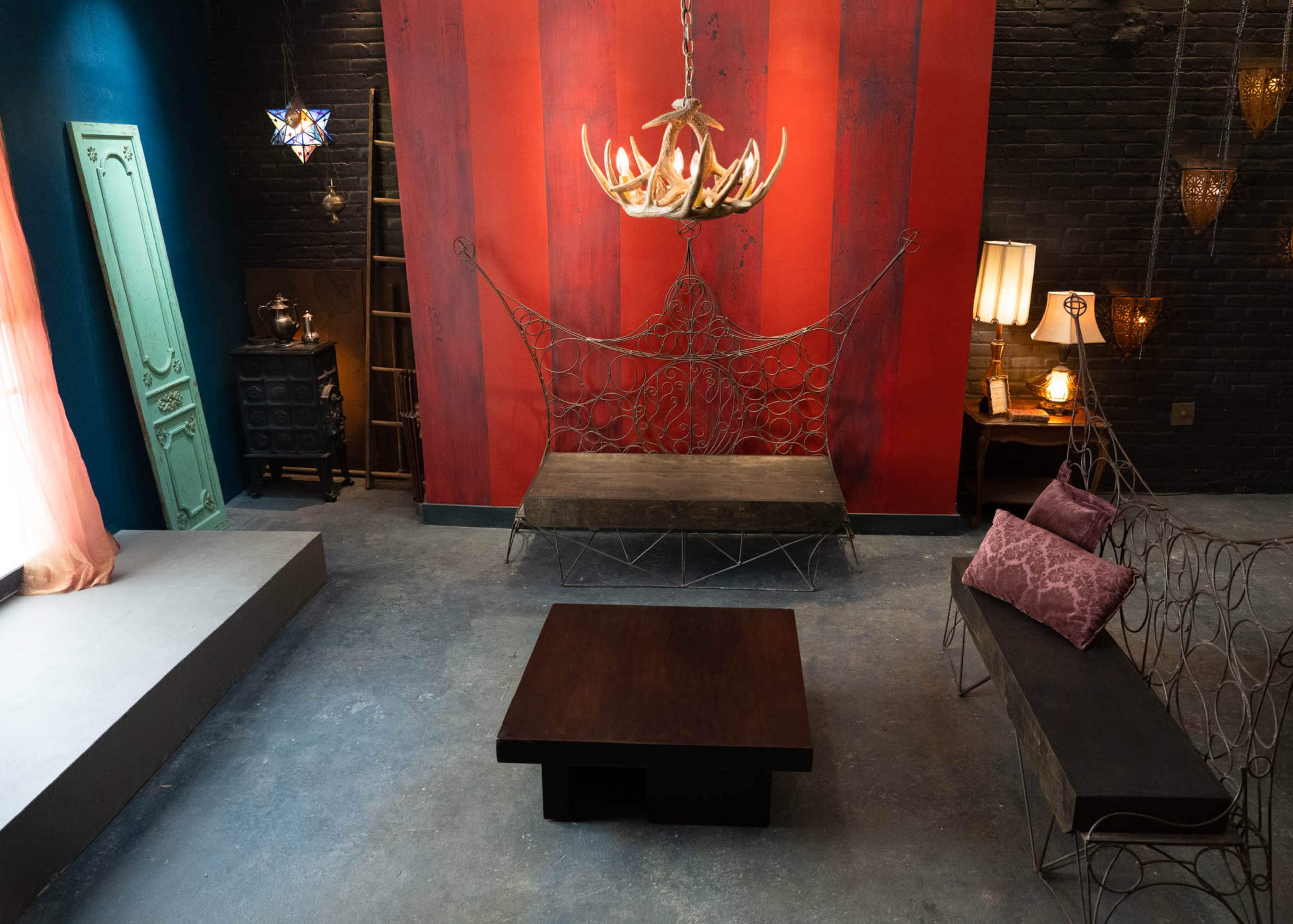 The scene features a dimly lit room with red and black striped walls, a wrought iron chandelier, two wrought iron benches, and a wooden coffee table at the center.