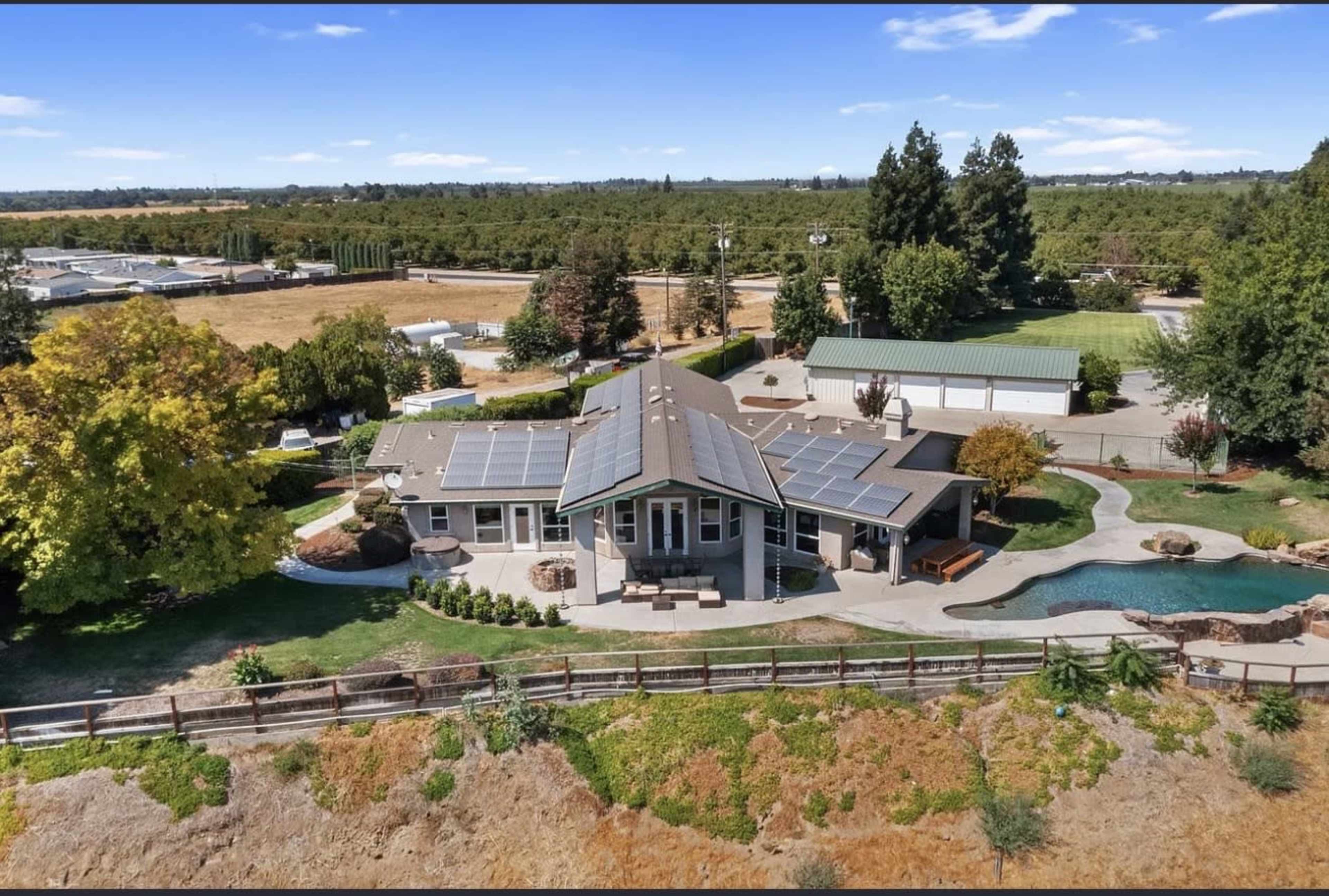 The image shows an aerial view of a single-story house with solar panels on the roof, surrounded by a landscaped yard and a swimming pool, set against a backdrop of fields and trees.