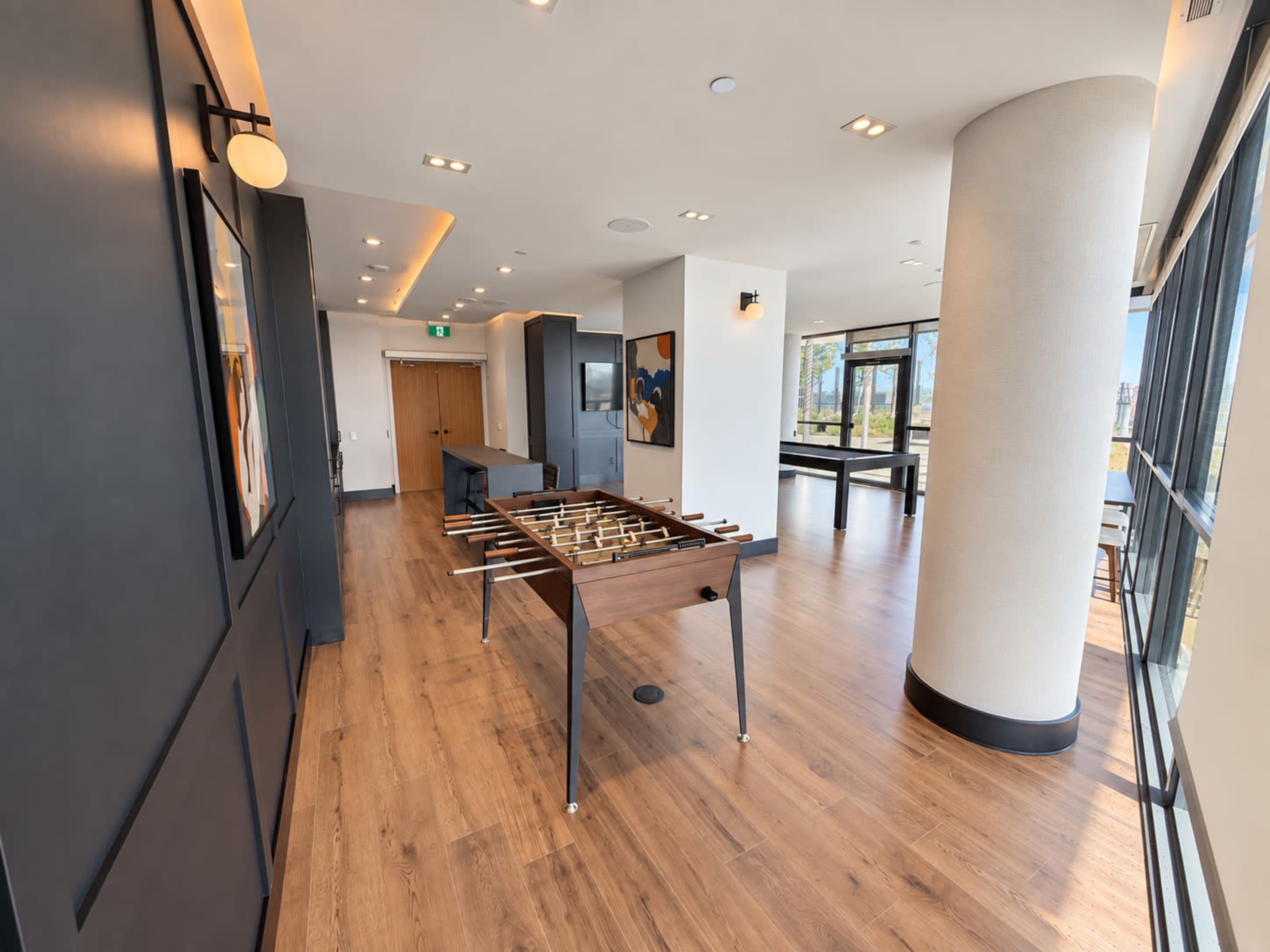 The image shows a modern recreational area with a foosball table, wooden flooring, and large windows letting in natural light.