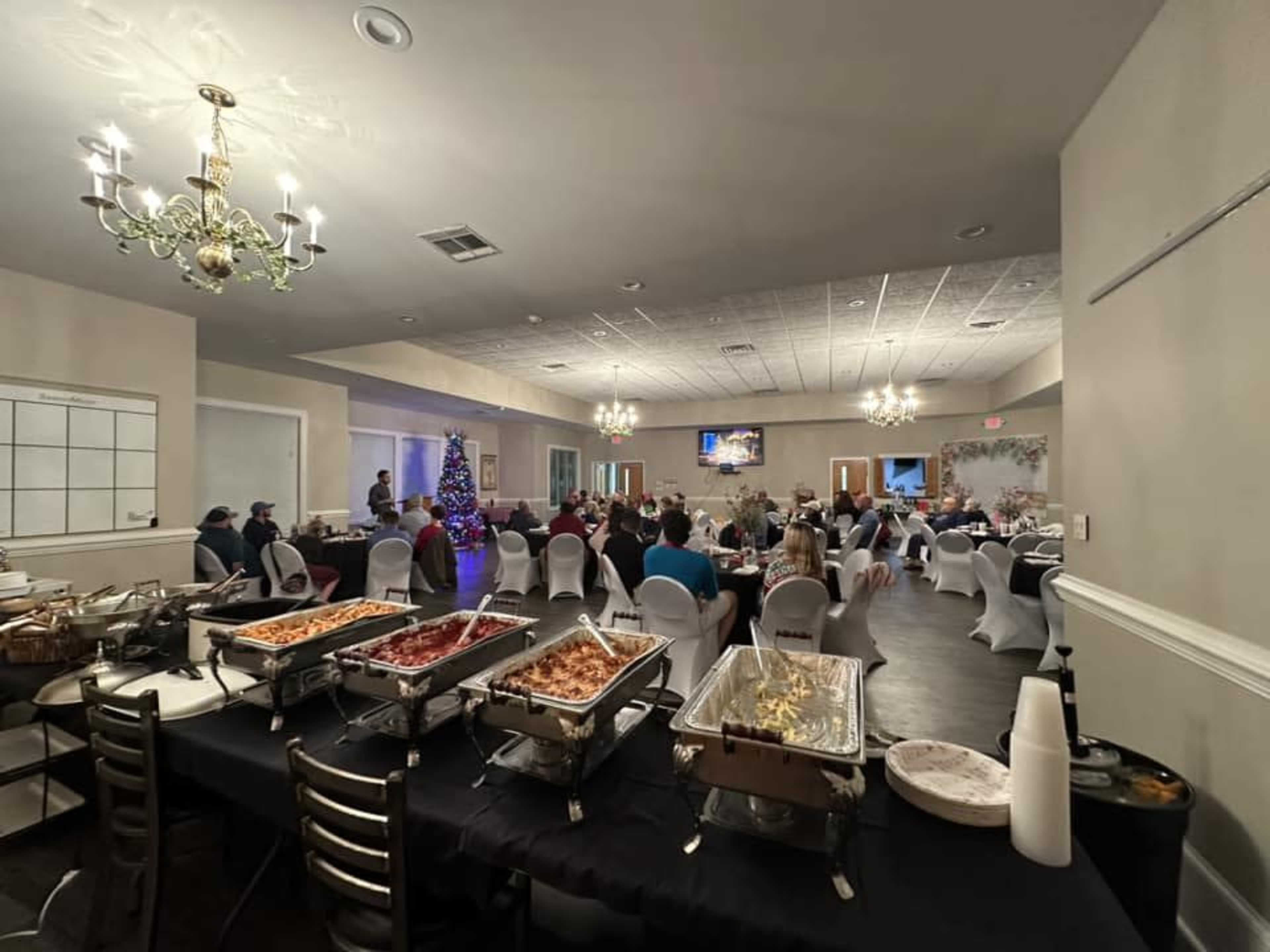 A banquet hall set up for an event, featuring white tablecloths, a buffet table with food, and guests seated at tables with a Christmas tree in the corner.