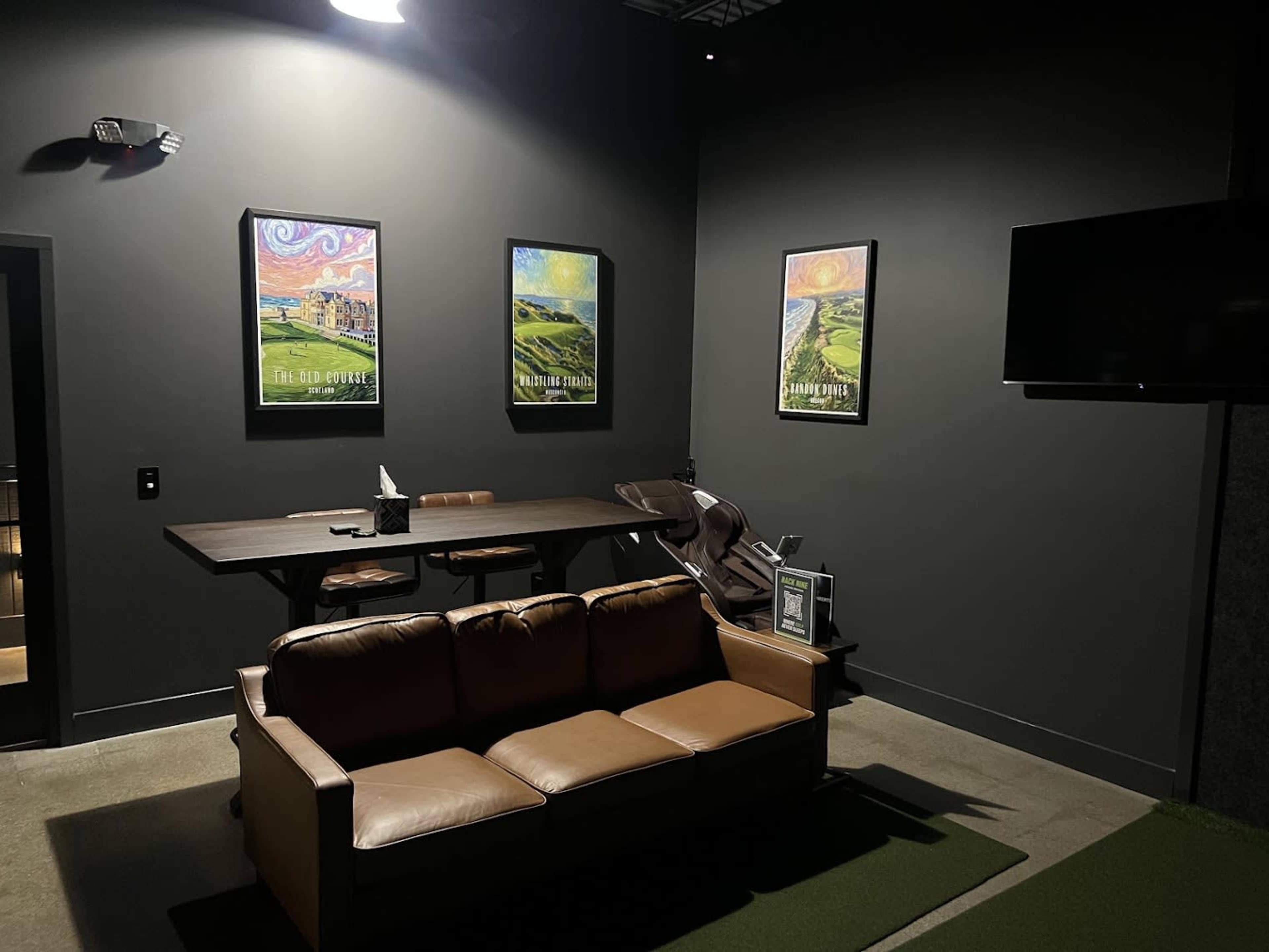 The image shows a dimly lit room featuring a brown leather couch, a wooden table with chairs, several framed colorful paintings on the wall, and a flat-screen TV.