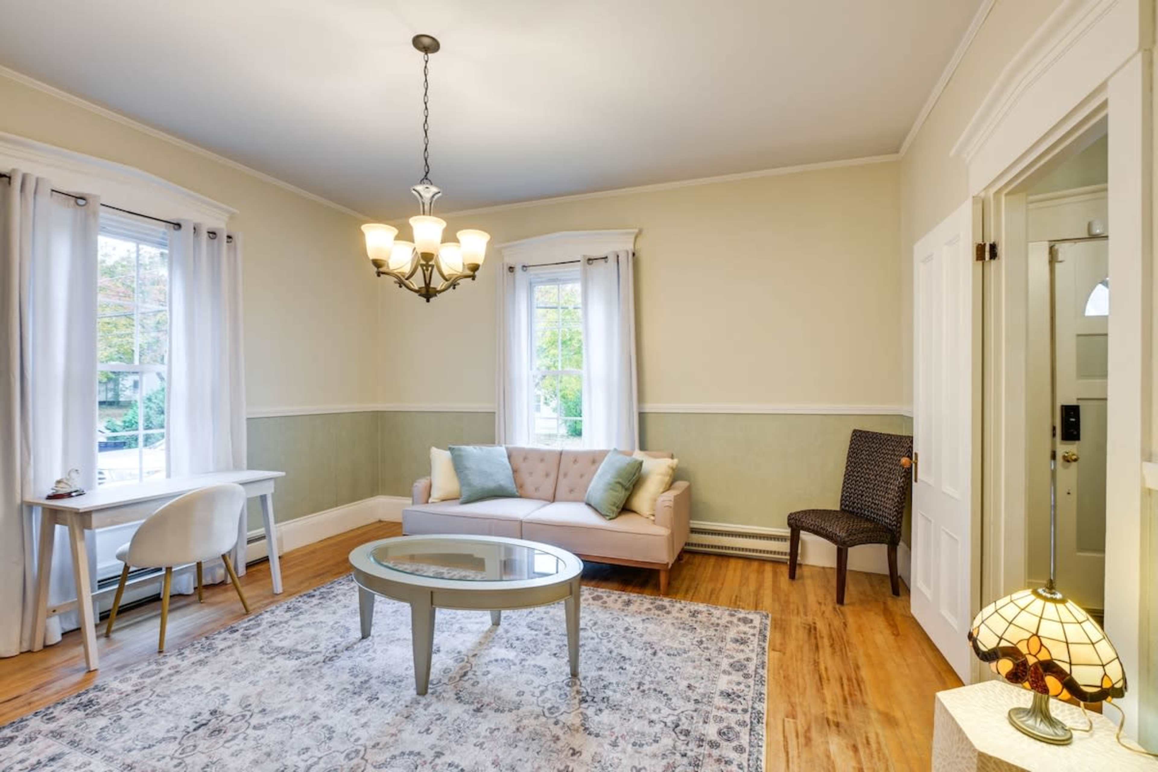 A softly lit living room featuring a beige sofa, a round coffee table, two windows, a small dining table, and a decorative lamp.