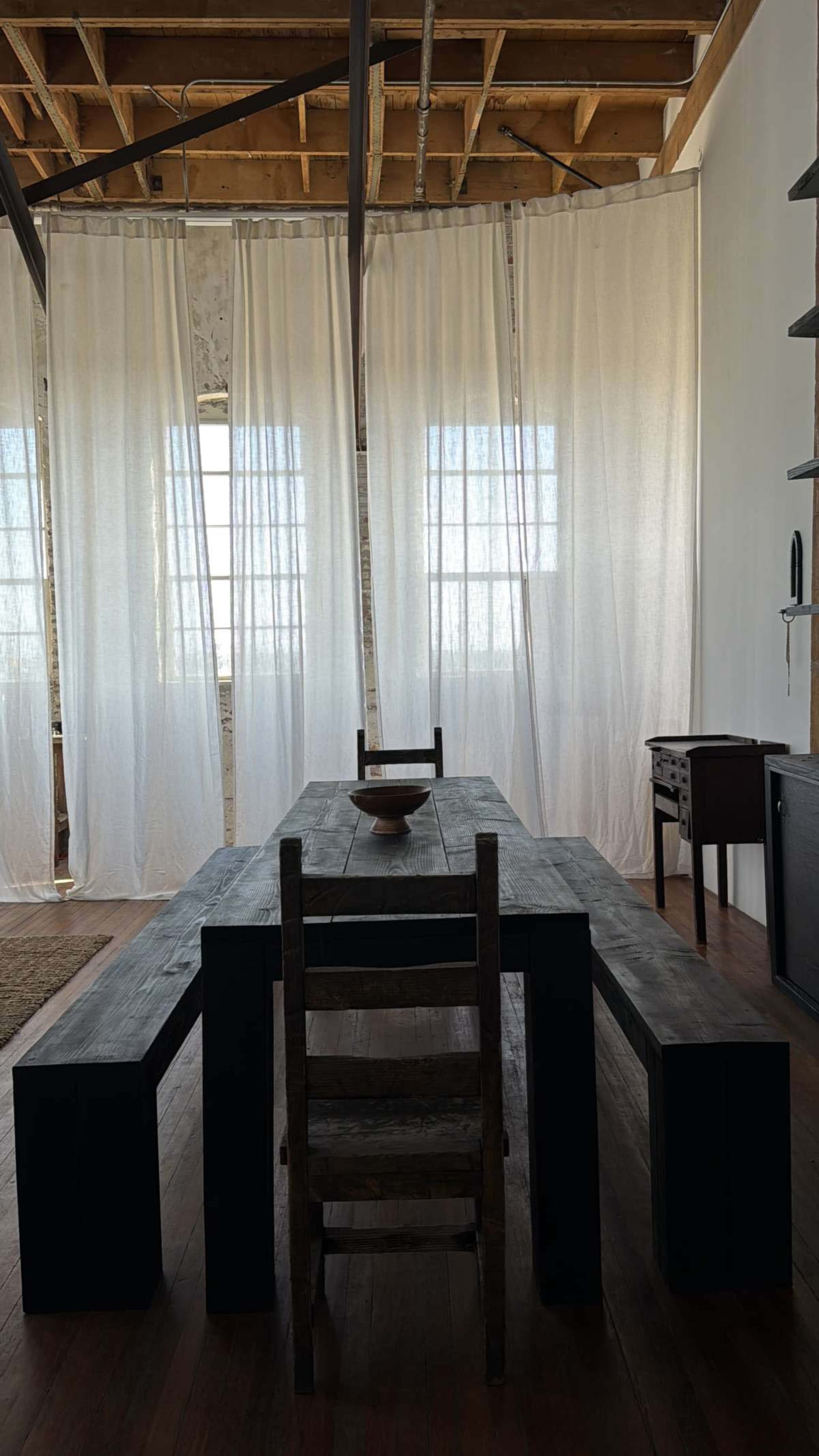 A rustic wooden table with benches and a single chair is positioned in a brightly lit room, separated by sheer curtains.
