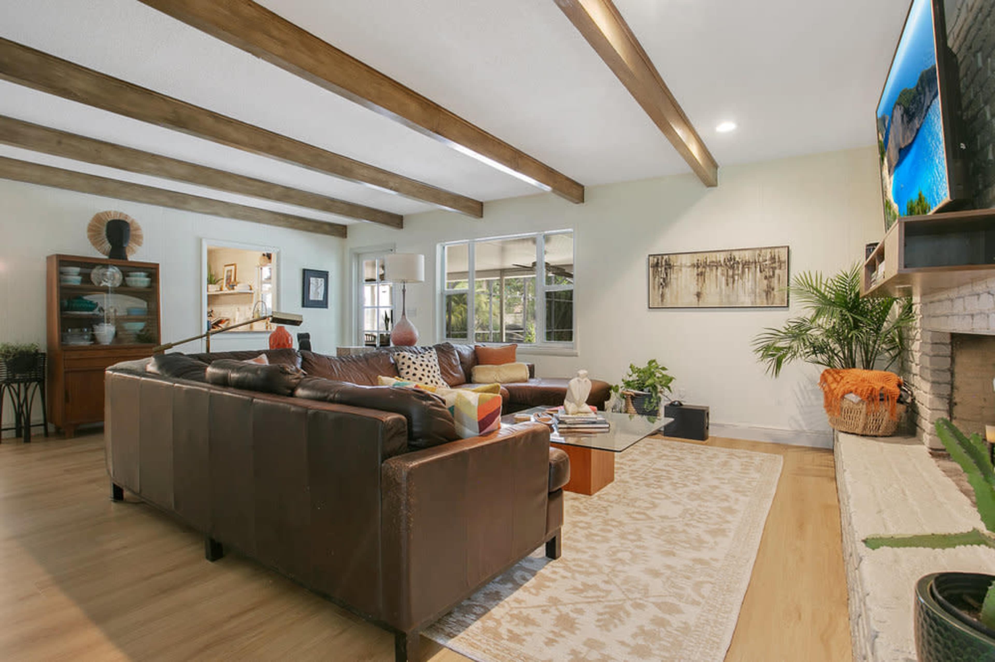 The image shows a spacious living room with brown leather sofas, wooden beams on the ceiling, a large TV mounted on the wall, and a decorative area rug on the floor.