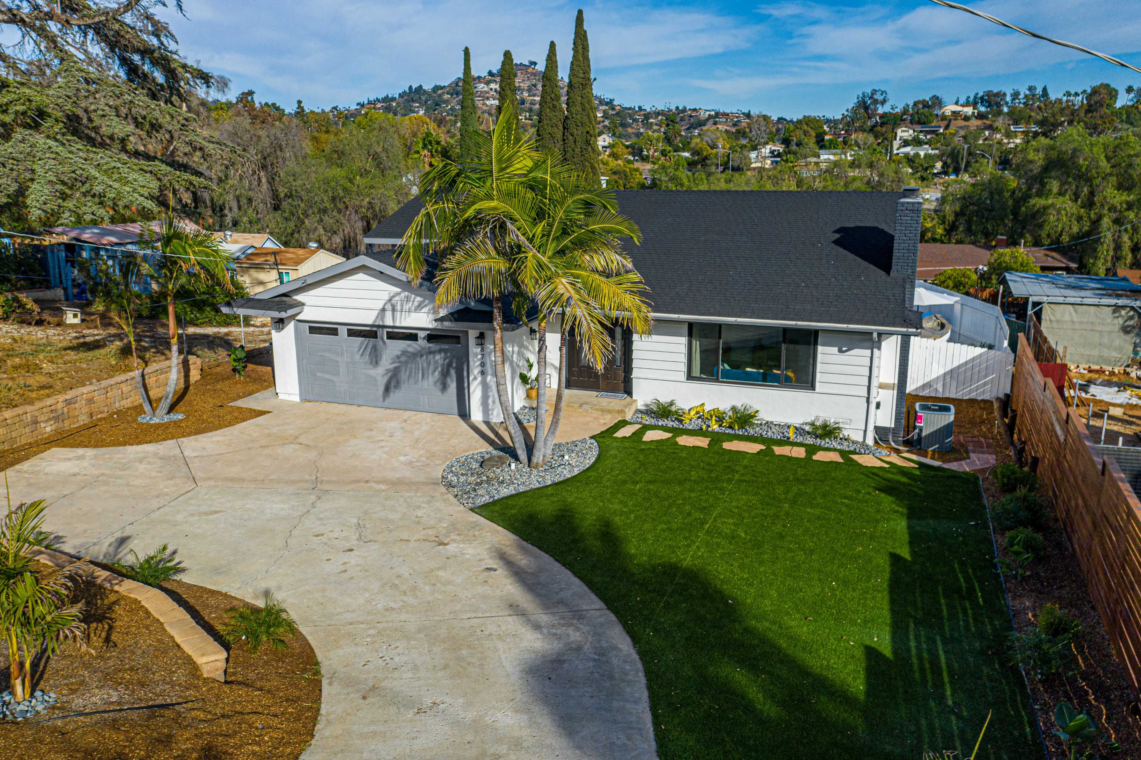 A modern single-family home with a two-car garage, a manicured lawn, and landscaping features including palm trees and a gravel path.