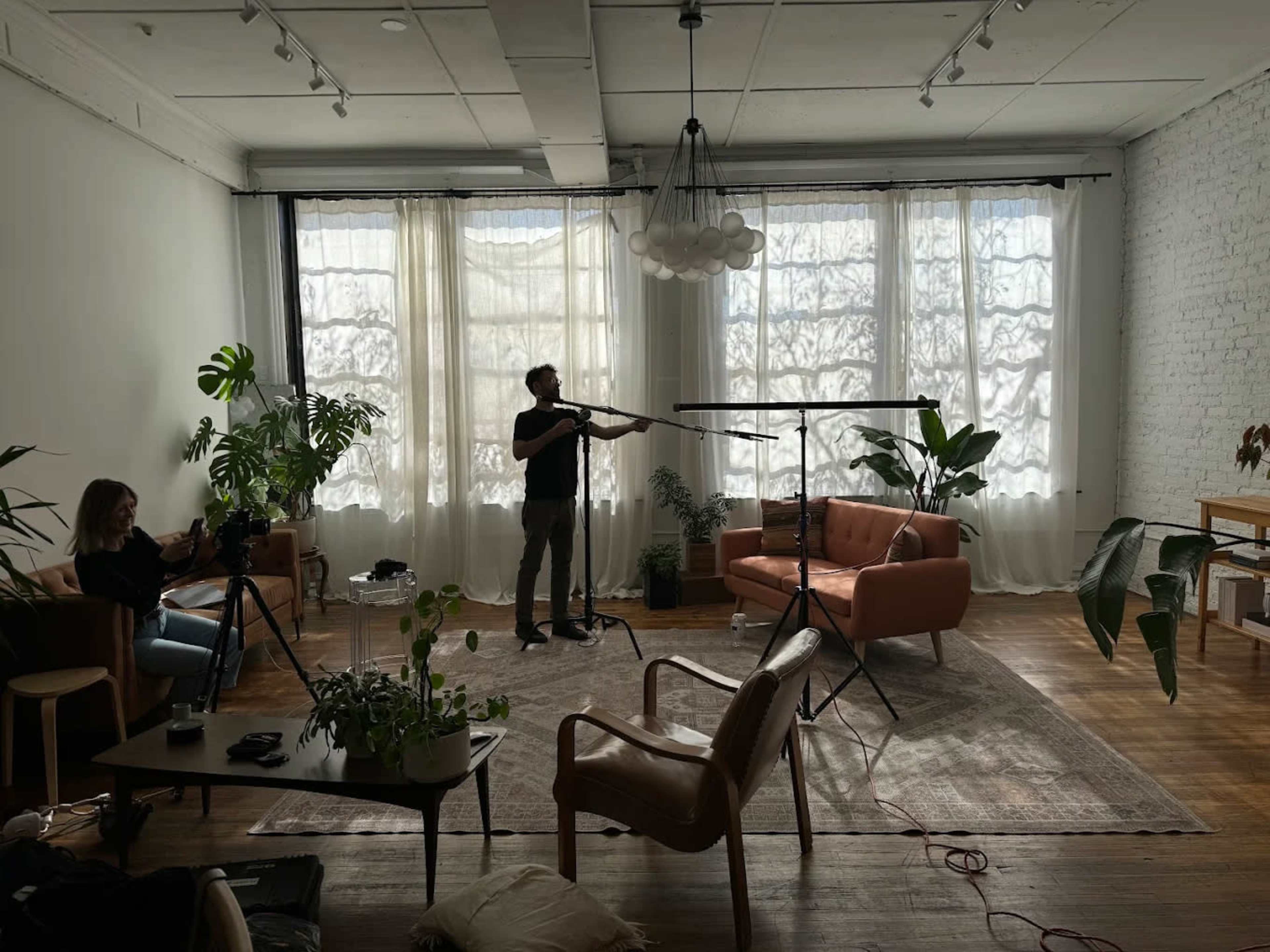 A person stands behind a microphone in a well-lit room with large windows, while another person sits nearby with a camera, surrounded by indoor plants and furniture.