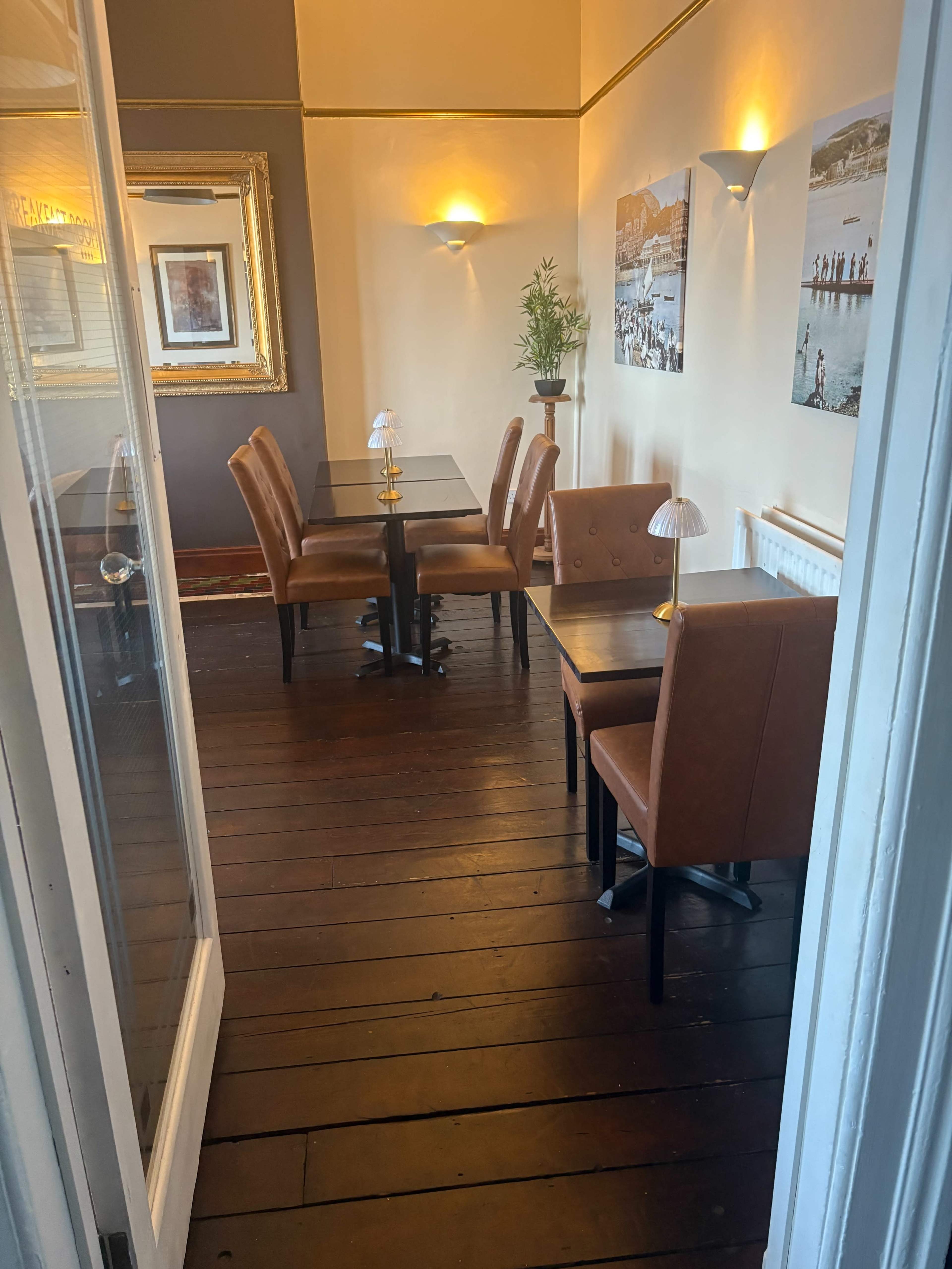The image shows a small dining area with two tables and brown leather chairs, featuring wooden flooring and wall decorations.