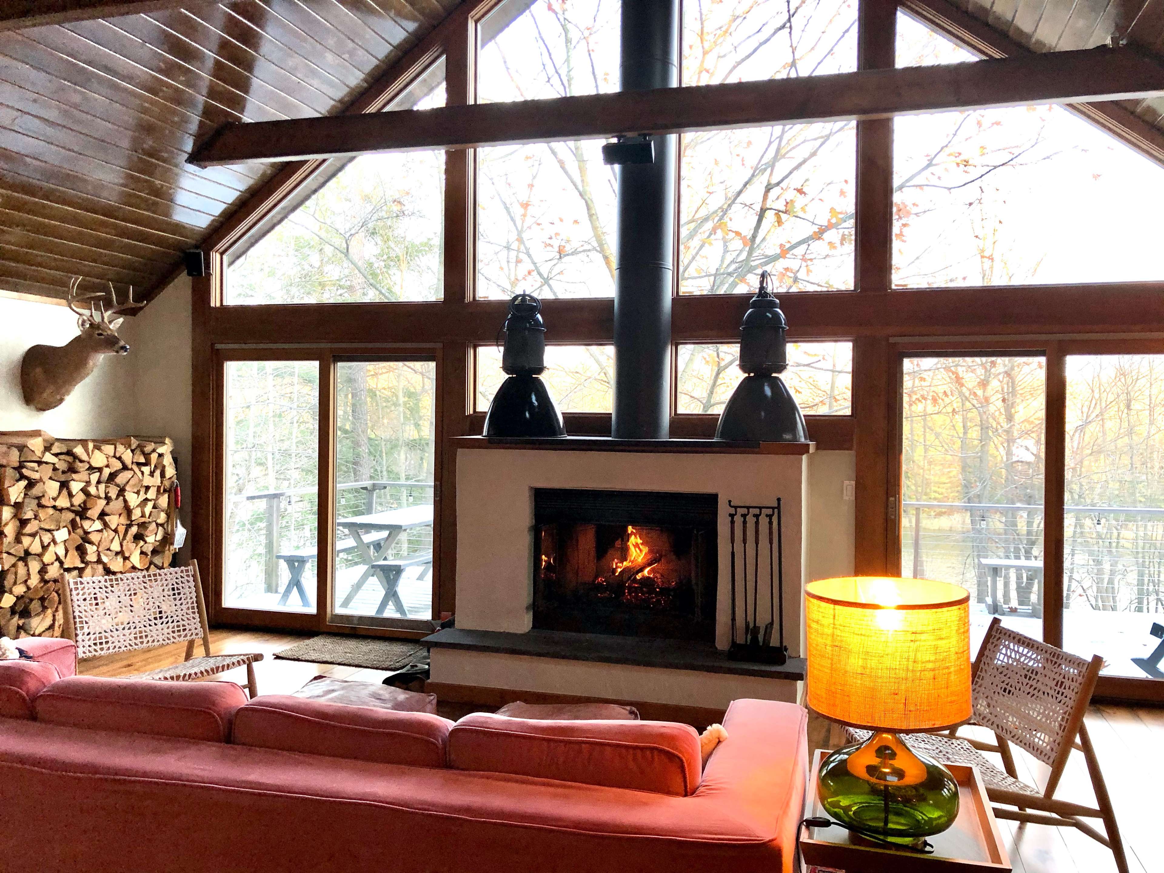 A cozy living room with a stone fireplace, large windows overlooking trees, and a woodpile beside the seating area.